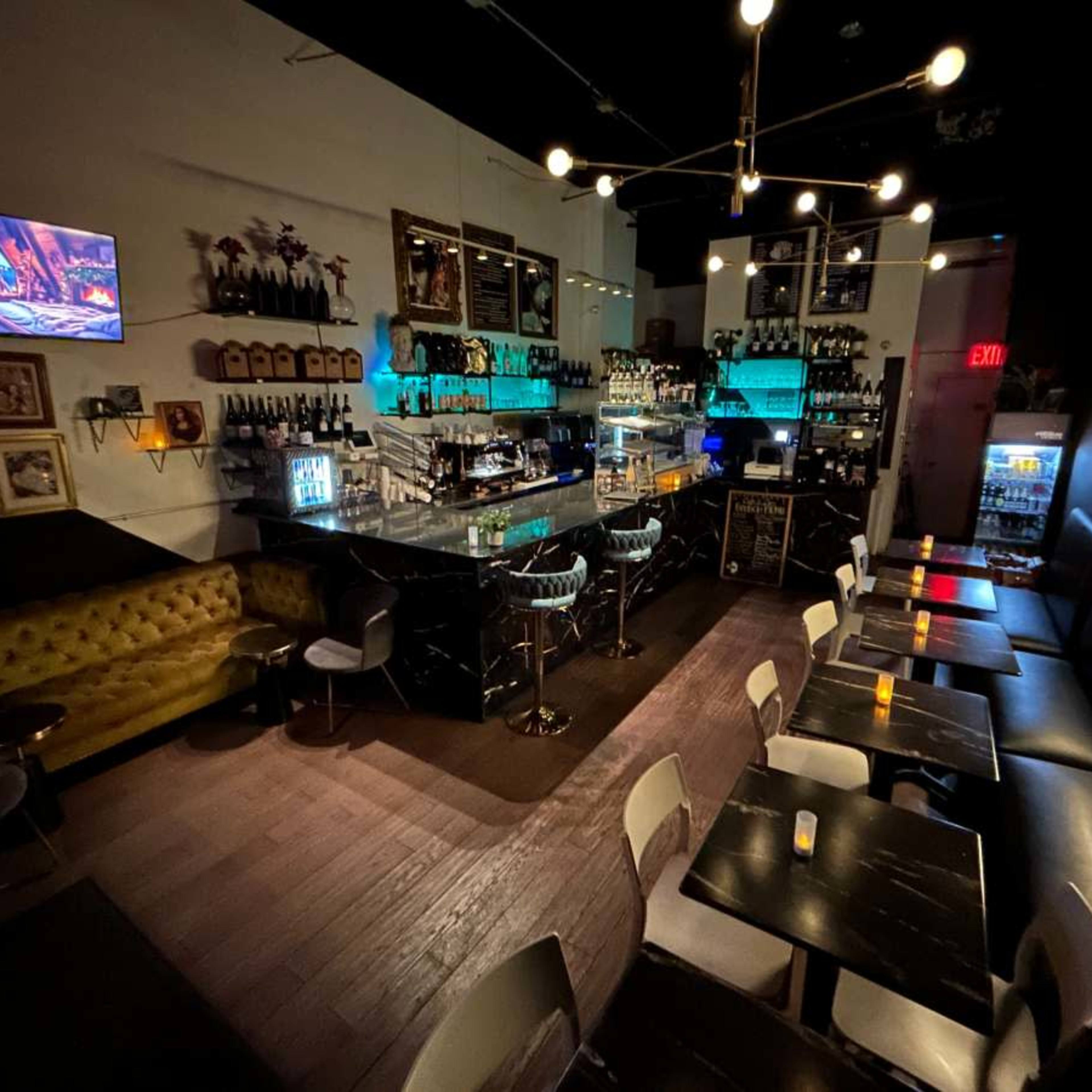 A dimly lit bar with a stylish counter, shelves filled with bottles, and seating areas featuring a mix of sofas and tables.