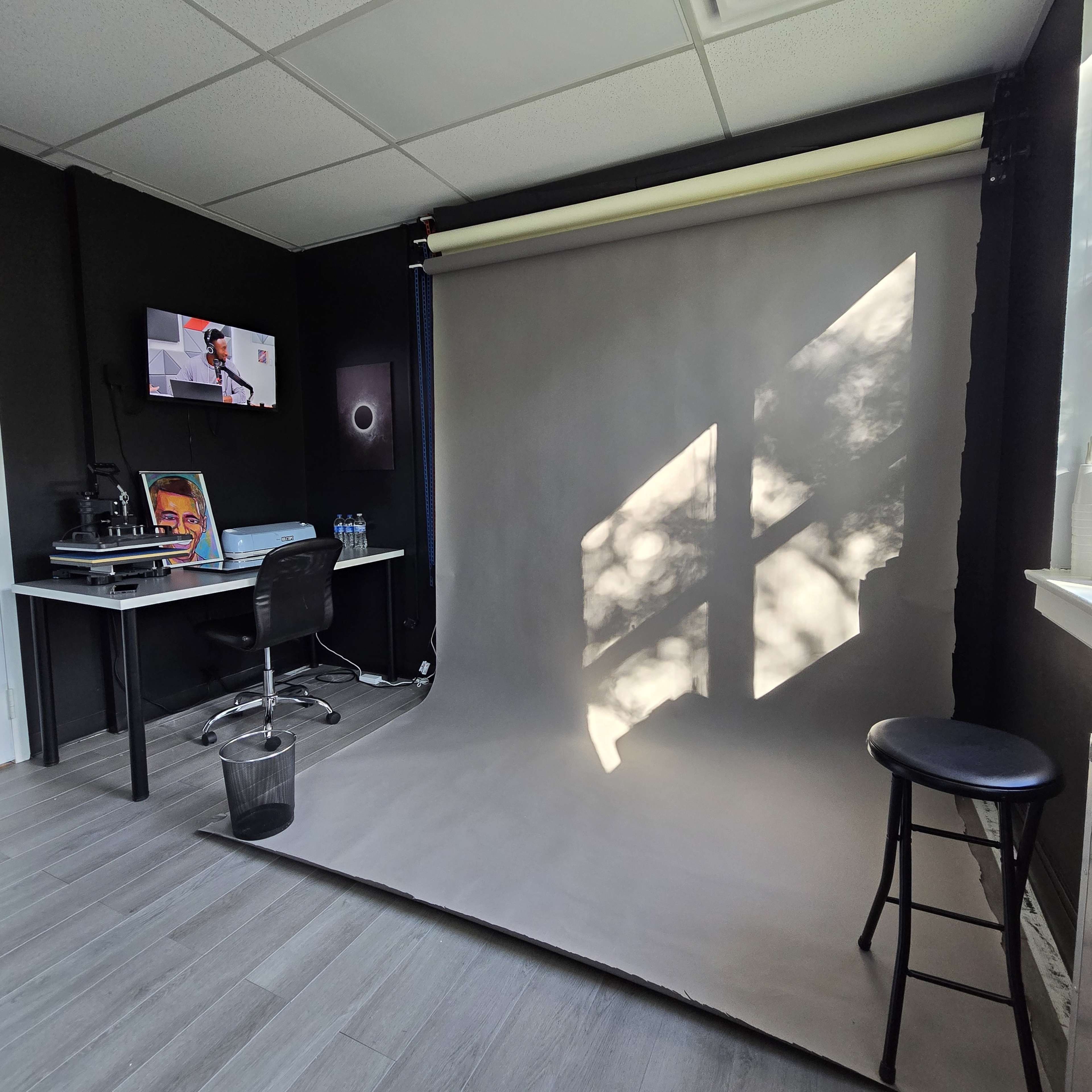A photography studio with a gray backdrop, a chair, a desk with a computer and monitor, and natural light casting shadows on the floor.