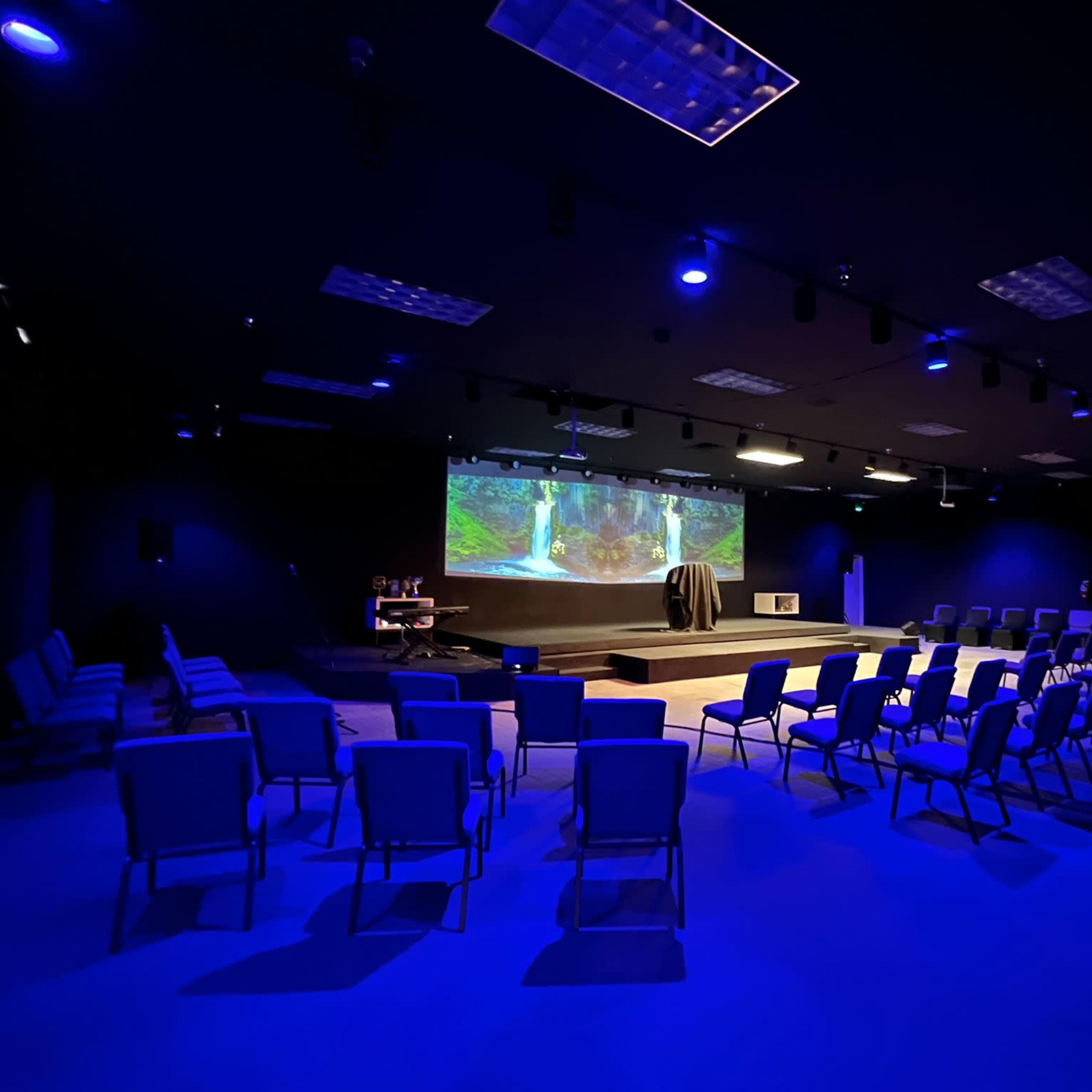 A dark room with blue lighting features rows of empty chairs facing a large projection screen displaying a nature scene.