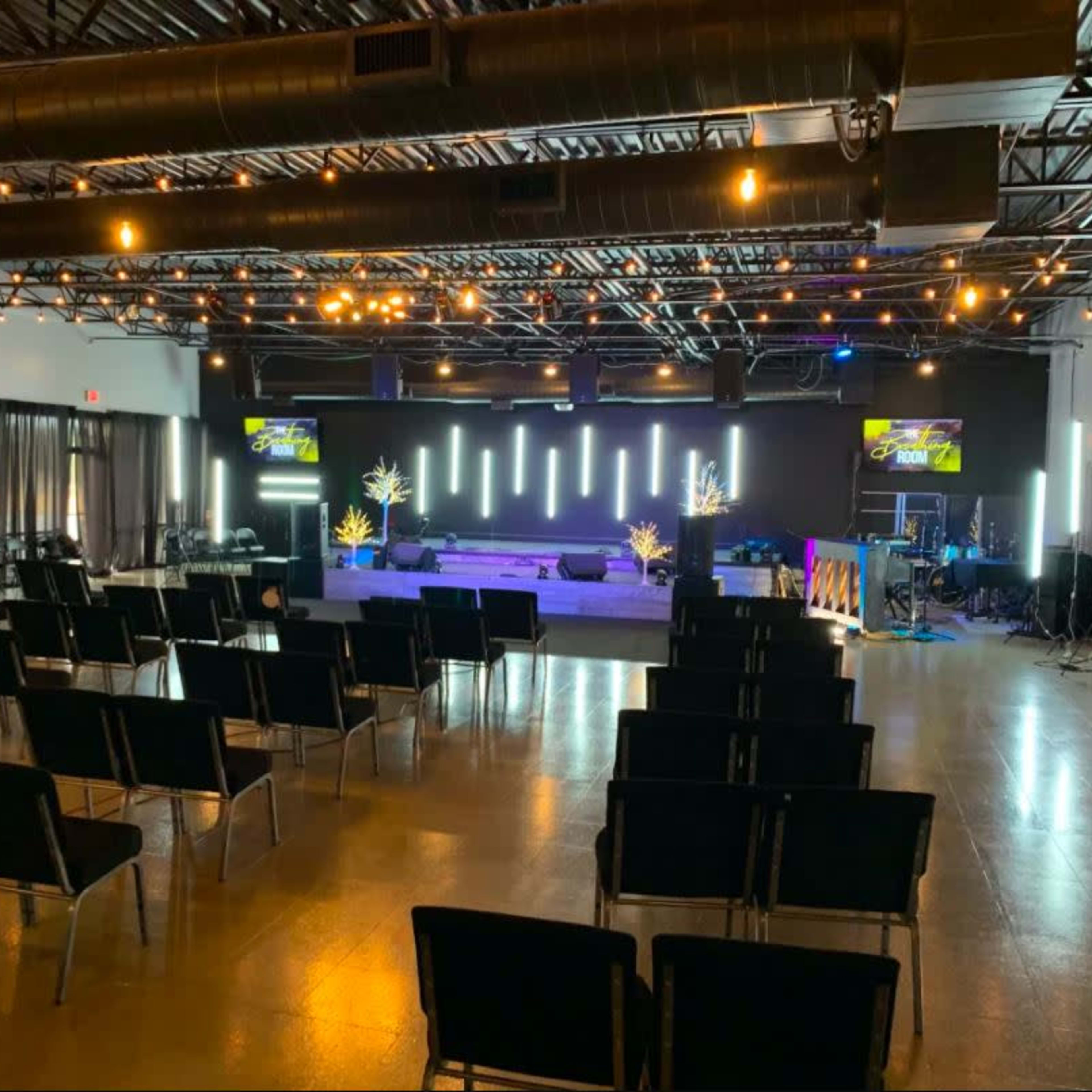 A spacious indoor venue set up for an event, featuring rows of black chairs facing a stage with lighting and audiovisual equipment.