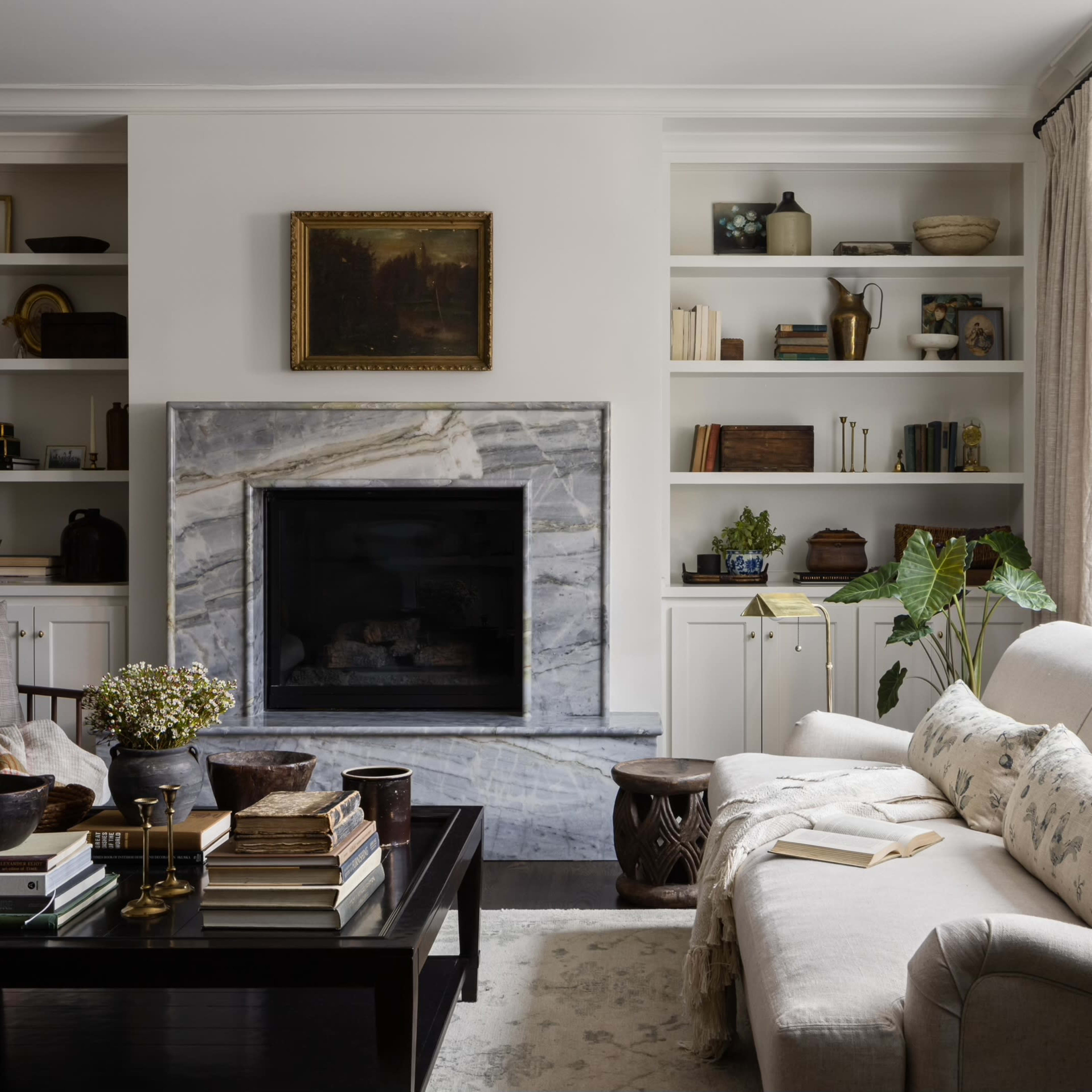 A tidy living room featuring a beige sofa, a marble fireplace, built-in shelves with various books and decor, and large windows allowing natural light.