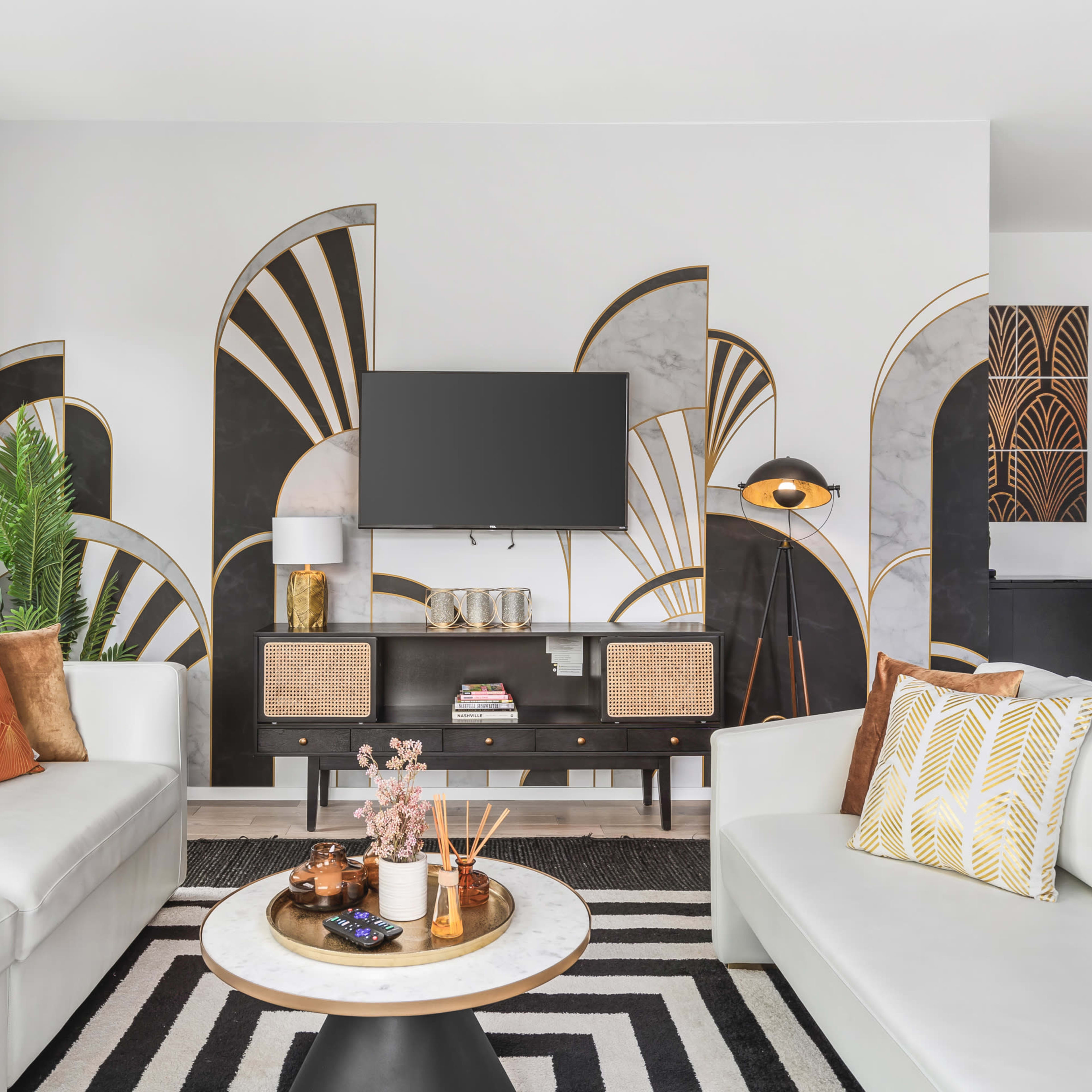 A modern living room features a black and white mural behind a stylish TV unit, with a round coffee table at the center and plush sofas adorned with orange and gold cushions.