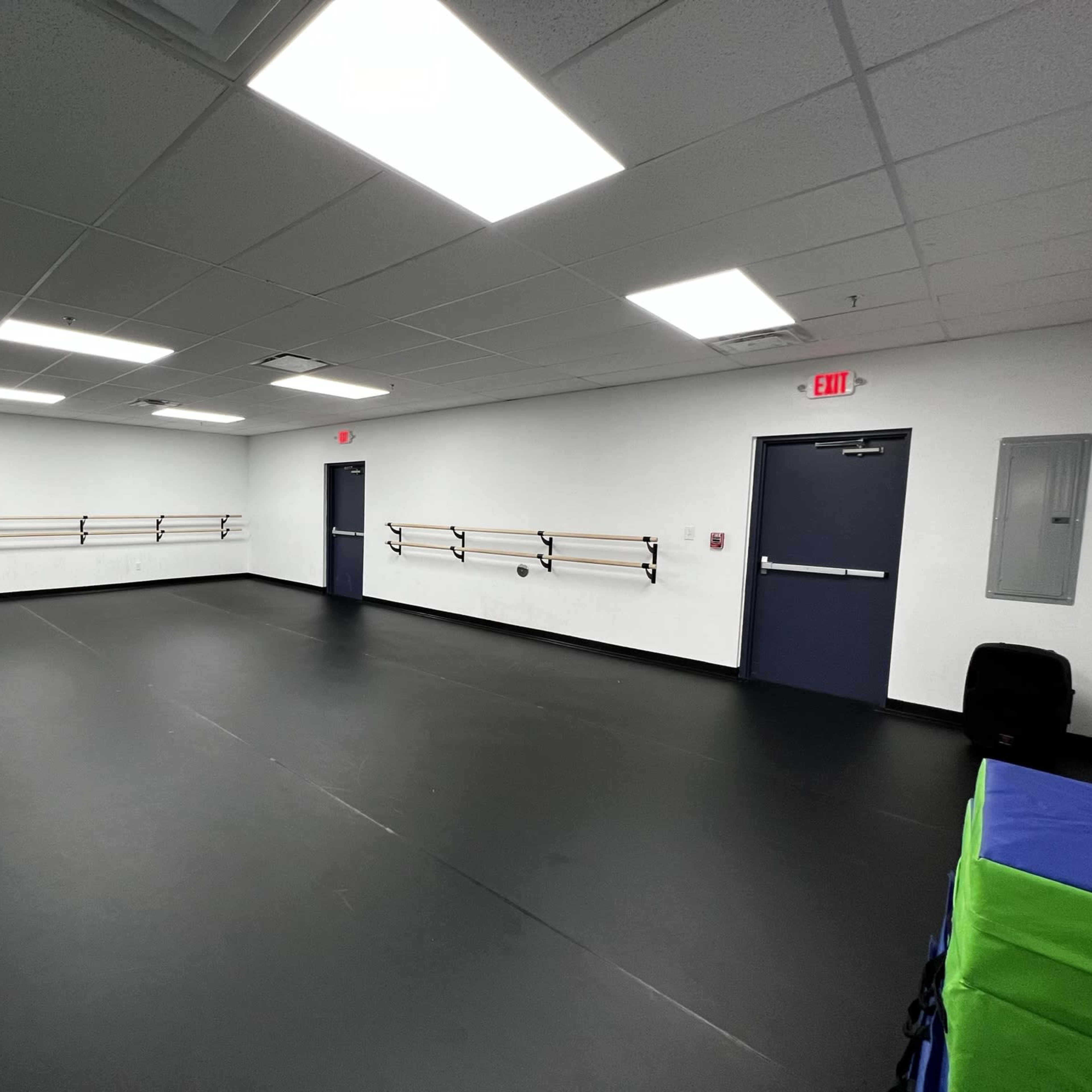 A spacious dance studio with black flooring, mirror walls, and wooden ballet bars mounted on the walls.