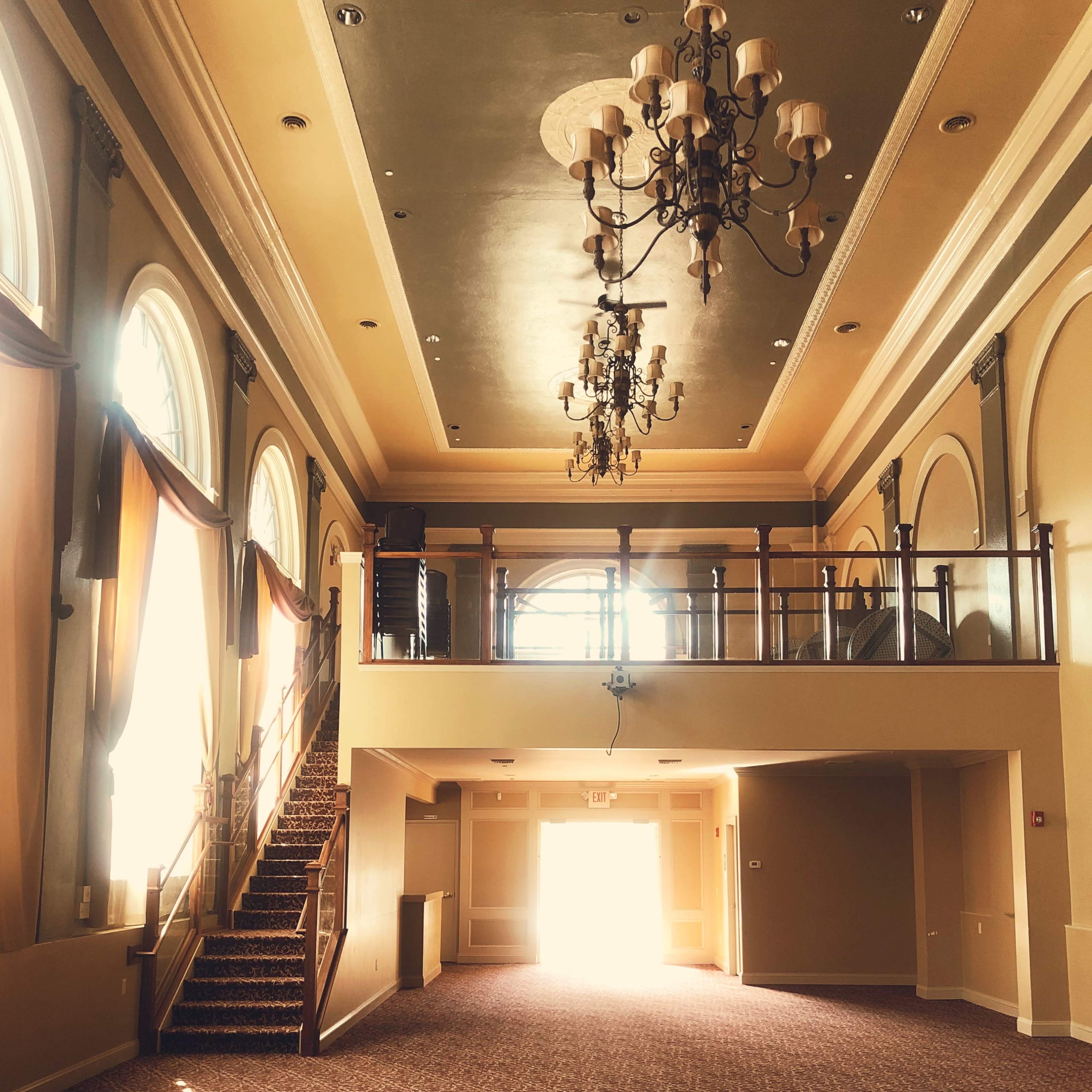 The spacious interior features large arched windows, a staircase leading to a balcony, and chandeliers hanging from a high ceiling.
