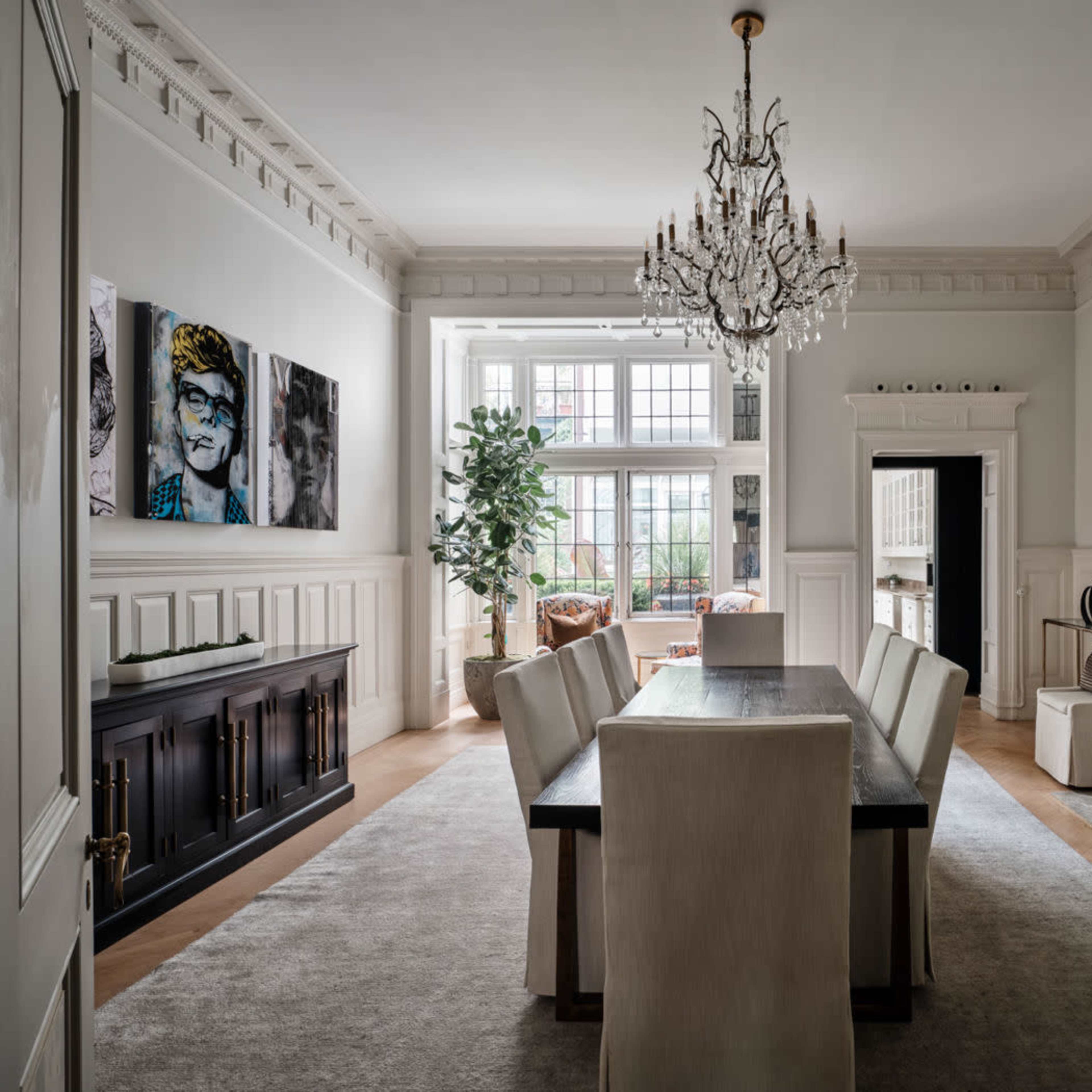 A spacious dining room featuring a long table surrounded by six upholstered chairs, a large chandelier overhead, and a decorative fireplace, all set against light-colored walls and wooden accents.