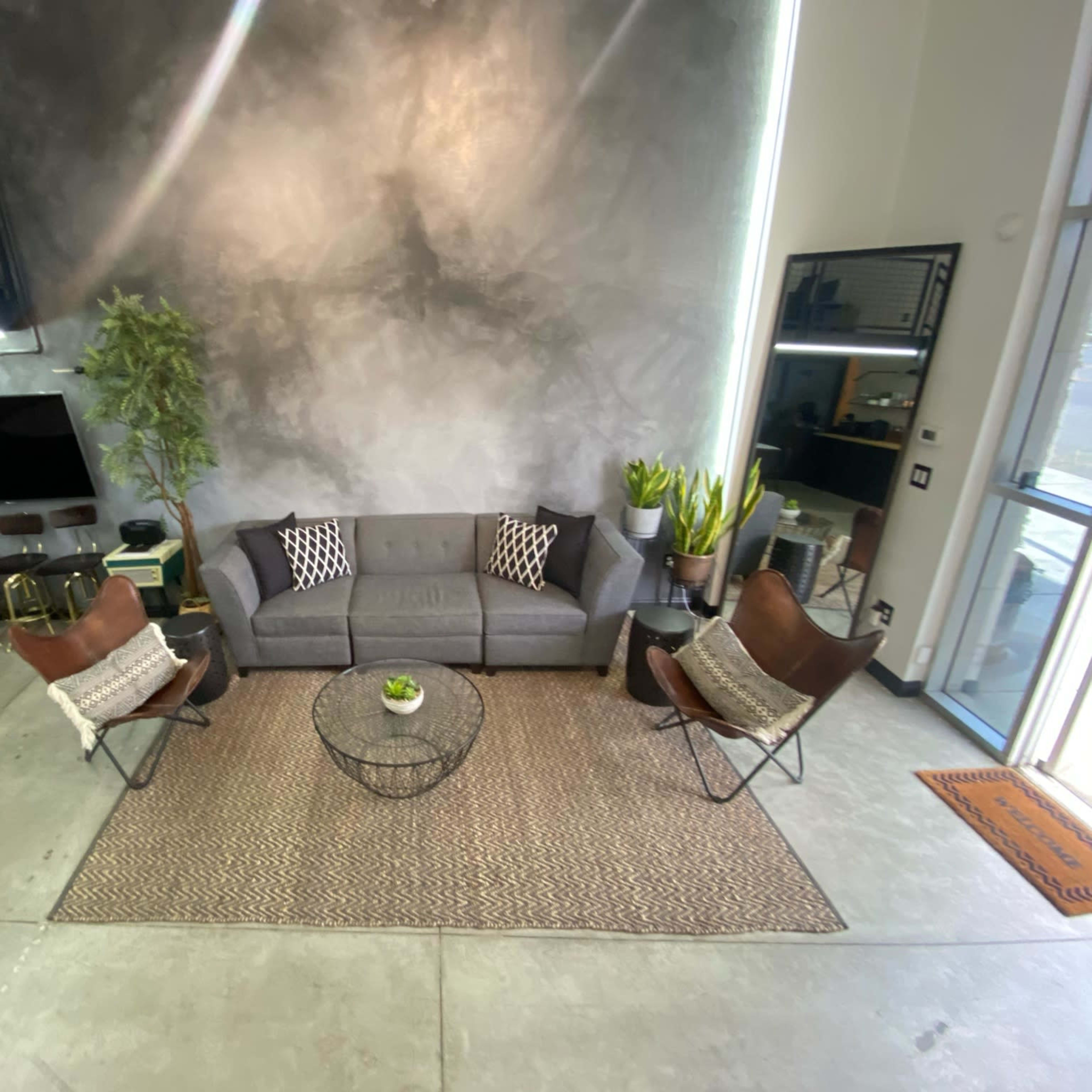 A modern living space with a gray sofa, two brown leather chairs, a round coffee table, and a rug, surrounded by plants and a large mirror.