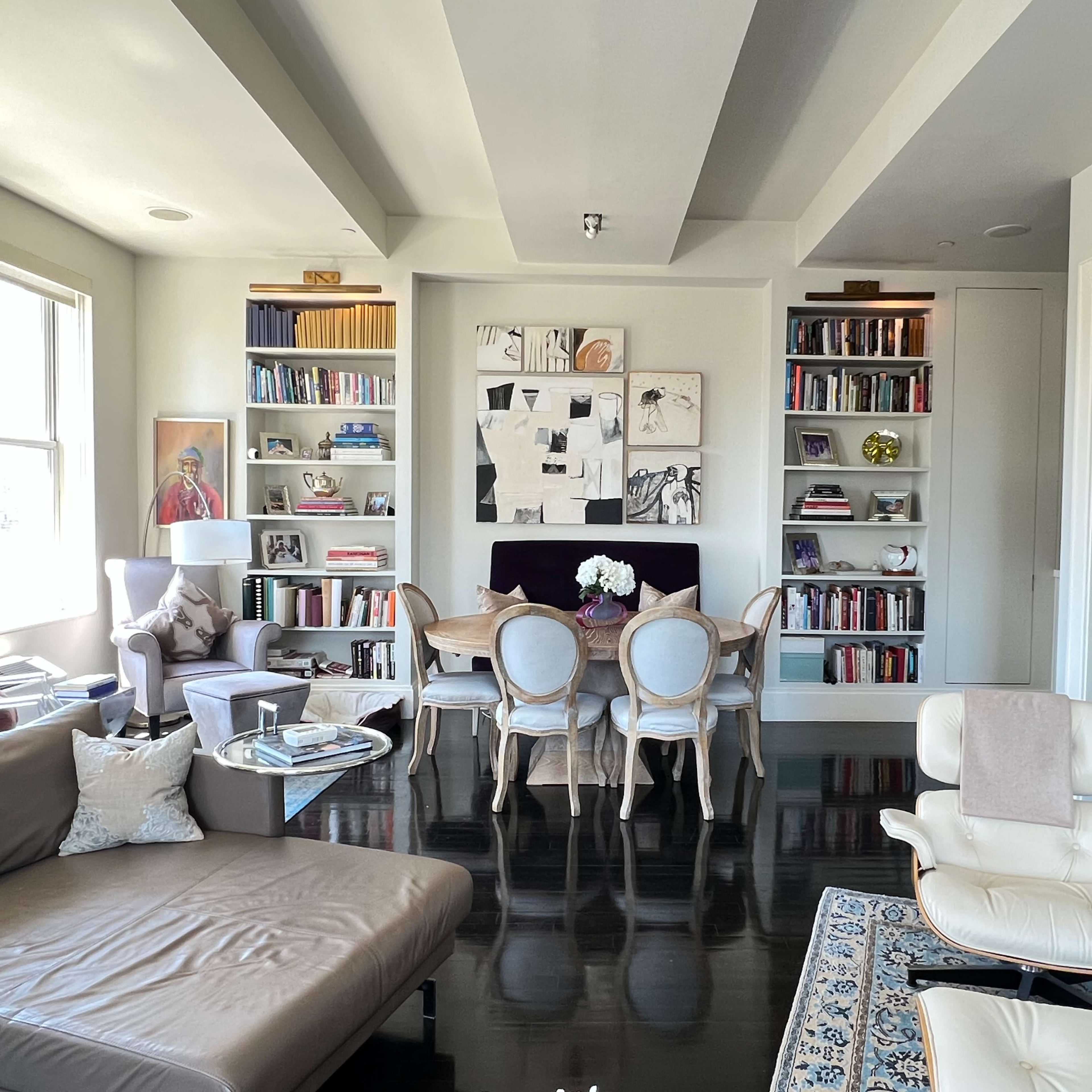 A modern living room features a large sofa, dining area with chairs and a table, bookshelves filled with books, and artwork on the walls.