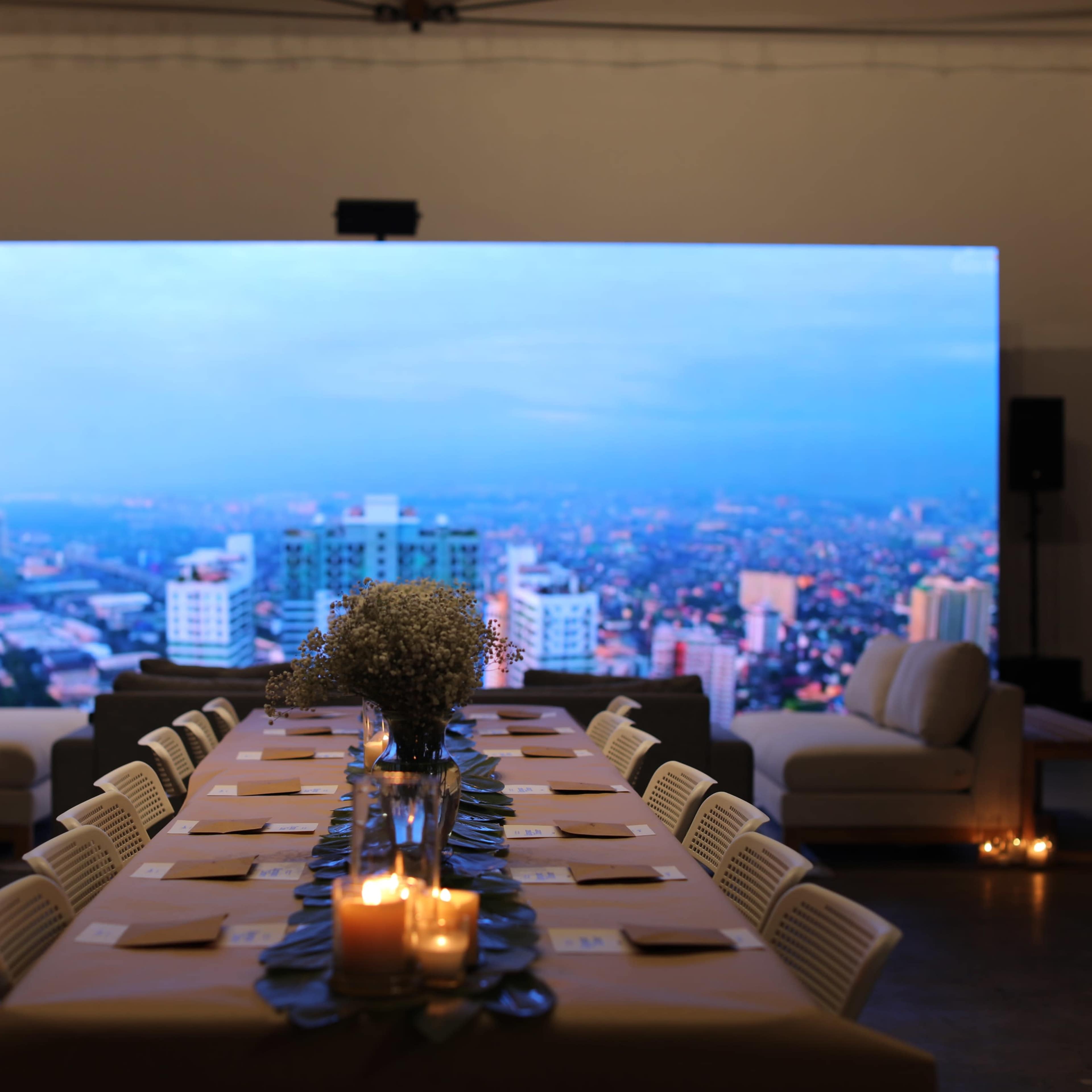 A long dining table is set in front of a large screen displaying a city skyline at dusk.