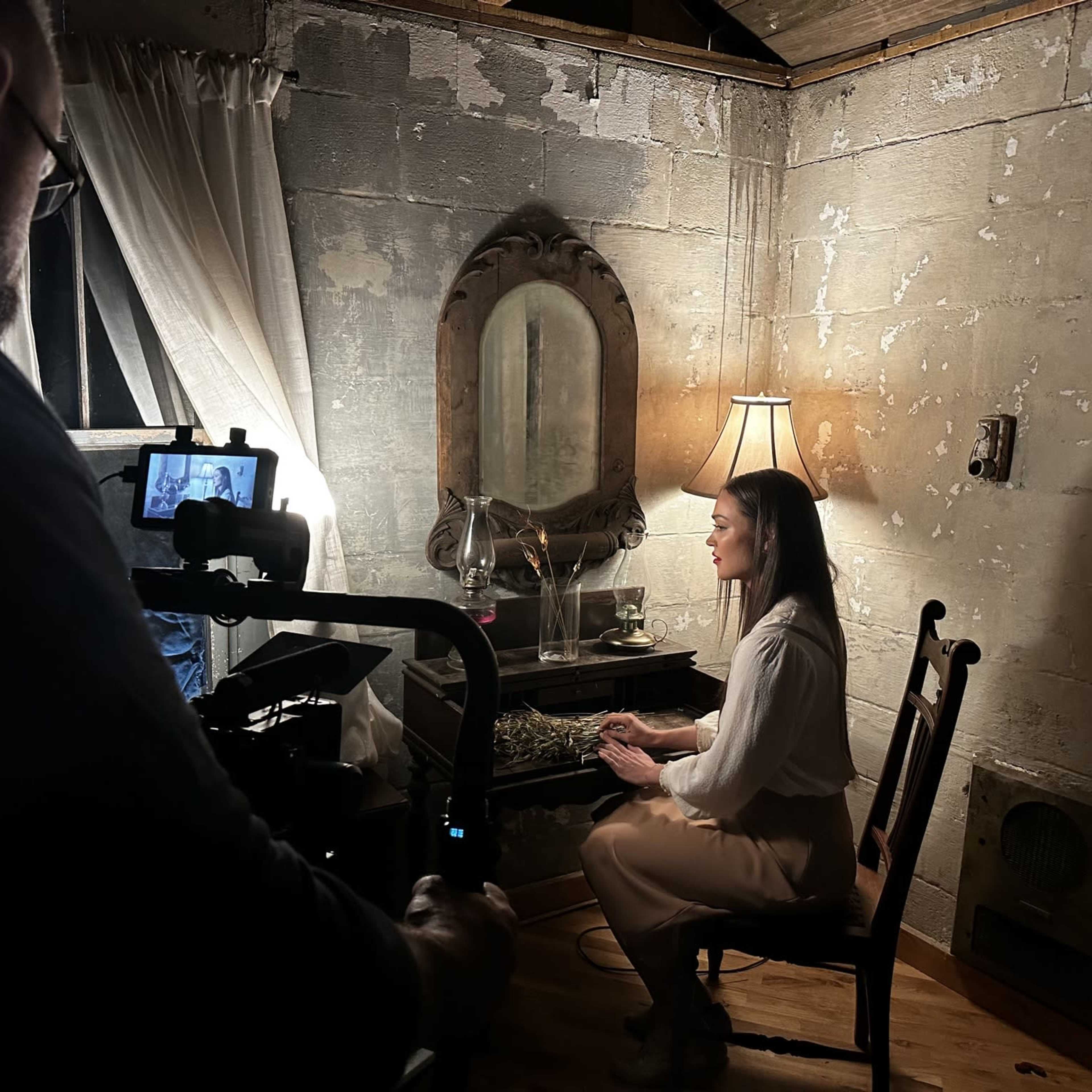 A person sits at a vanity table with a mirror, while another individual holds a camera and records the scene in a dimly lit room.