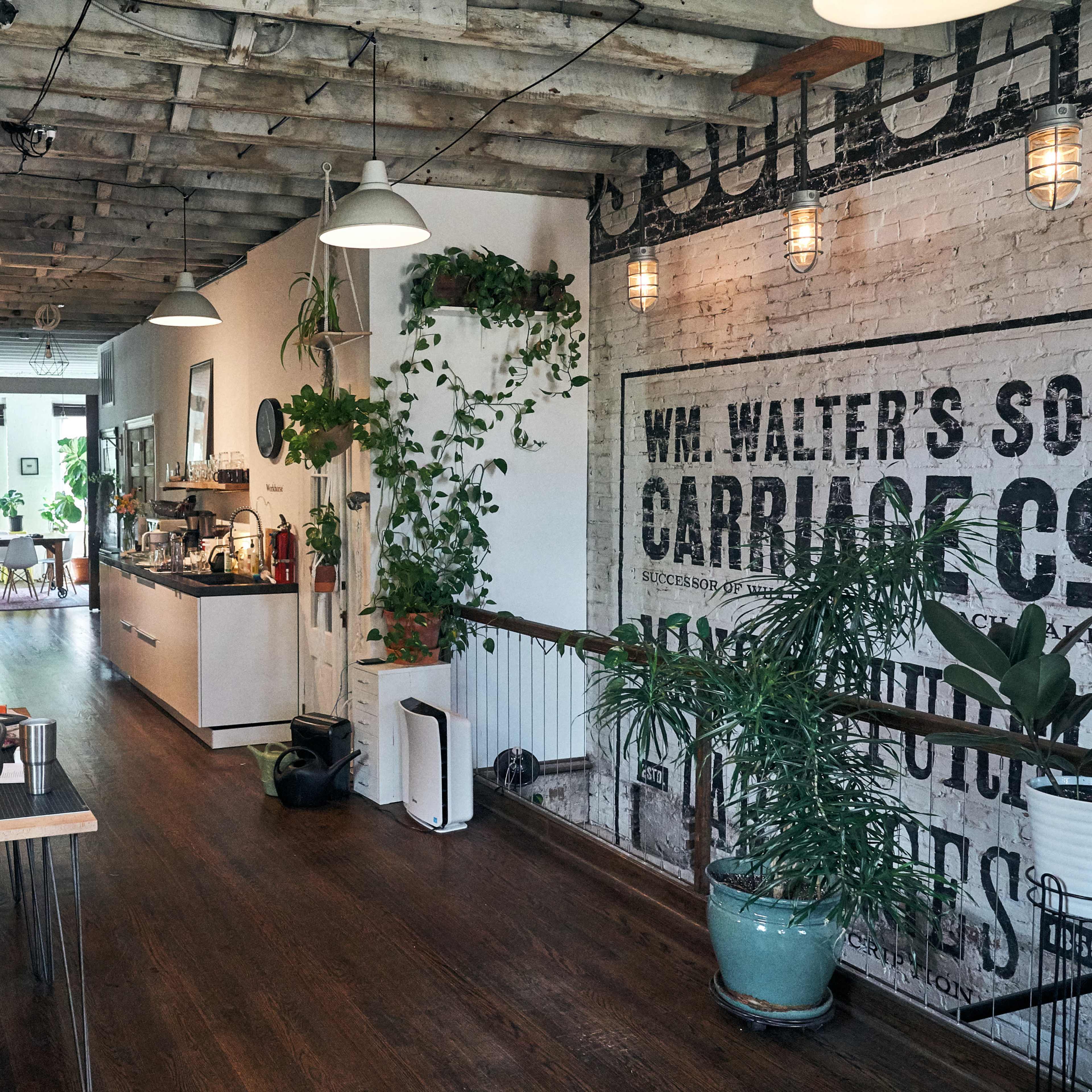 A spacious office interior with wooden beams, exposed brick walls, and plants, featuring workstations with two individuals seated at desks and a kitchenette area in the background.