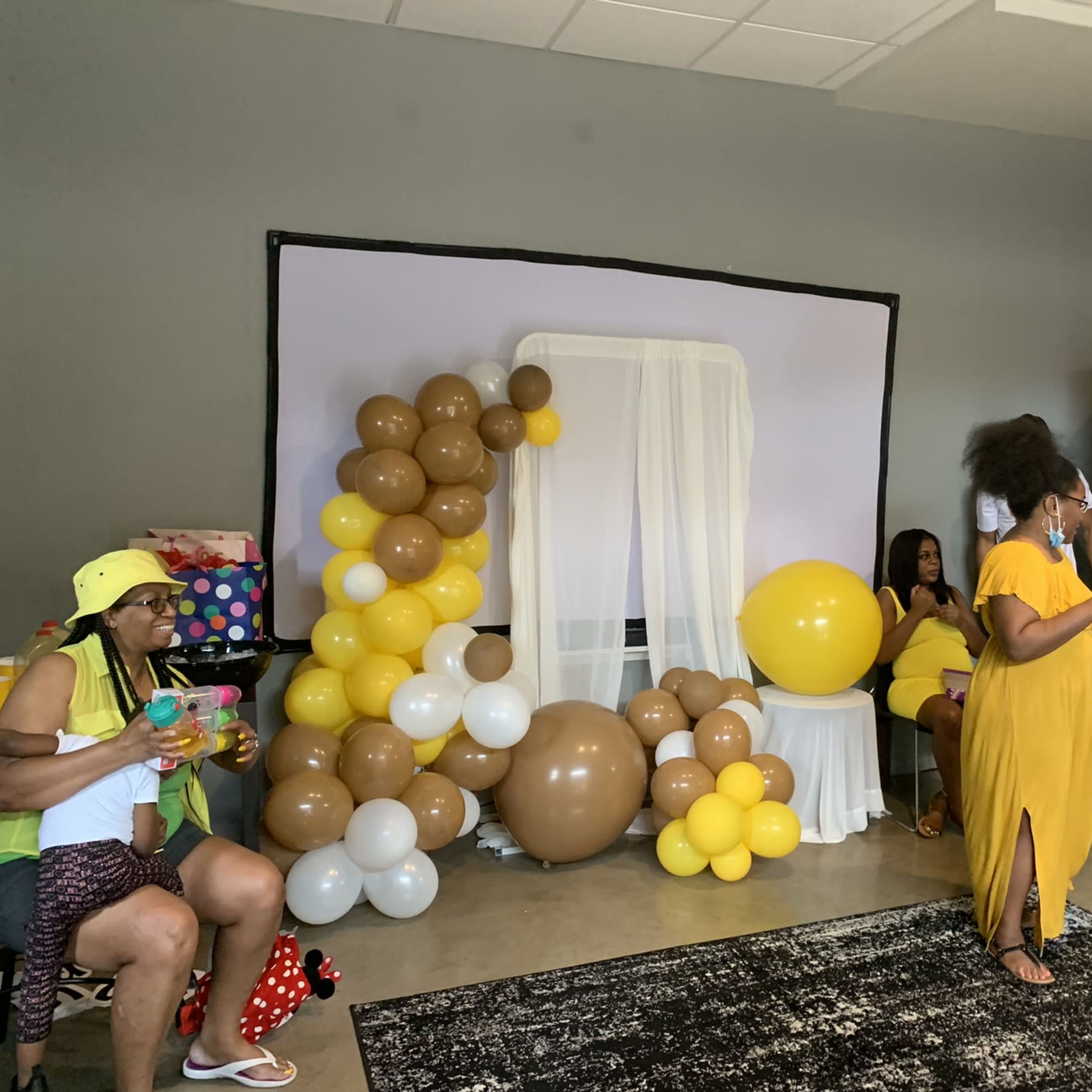 A gathering space features yellow and brown balloons arranged near a white backdrop, with guests seated and mingling at tables.