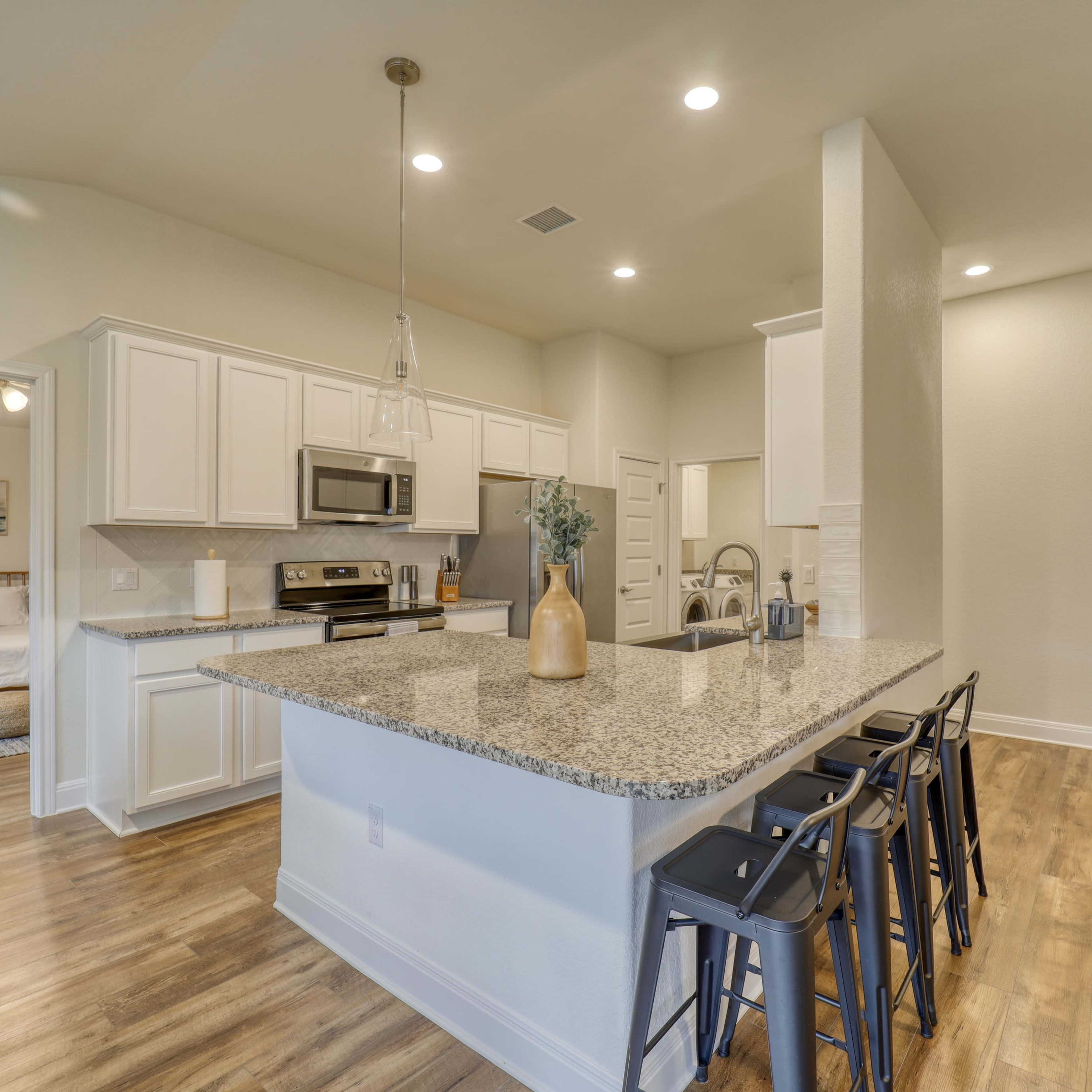 A modern kitchen features a large island with bar seating, white cabinetry, stainless steel appliances, and hardwood flooring, opening to a cozy bedroom area.
