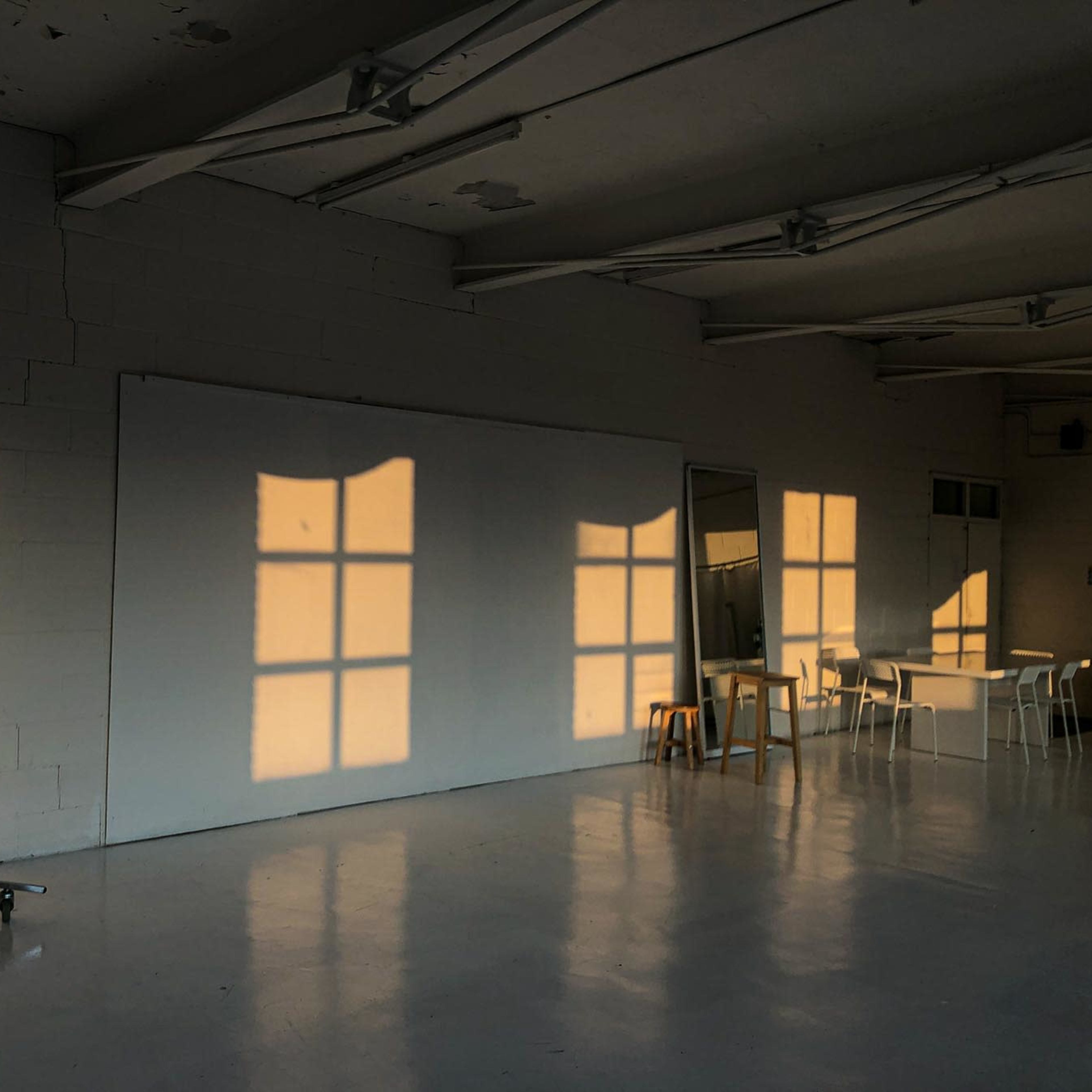 Shadows of a window and furniture are cast on a white wall in a sparsely furnished room with a polished floor.