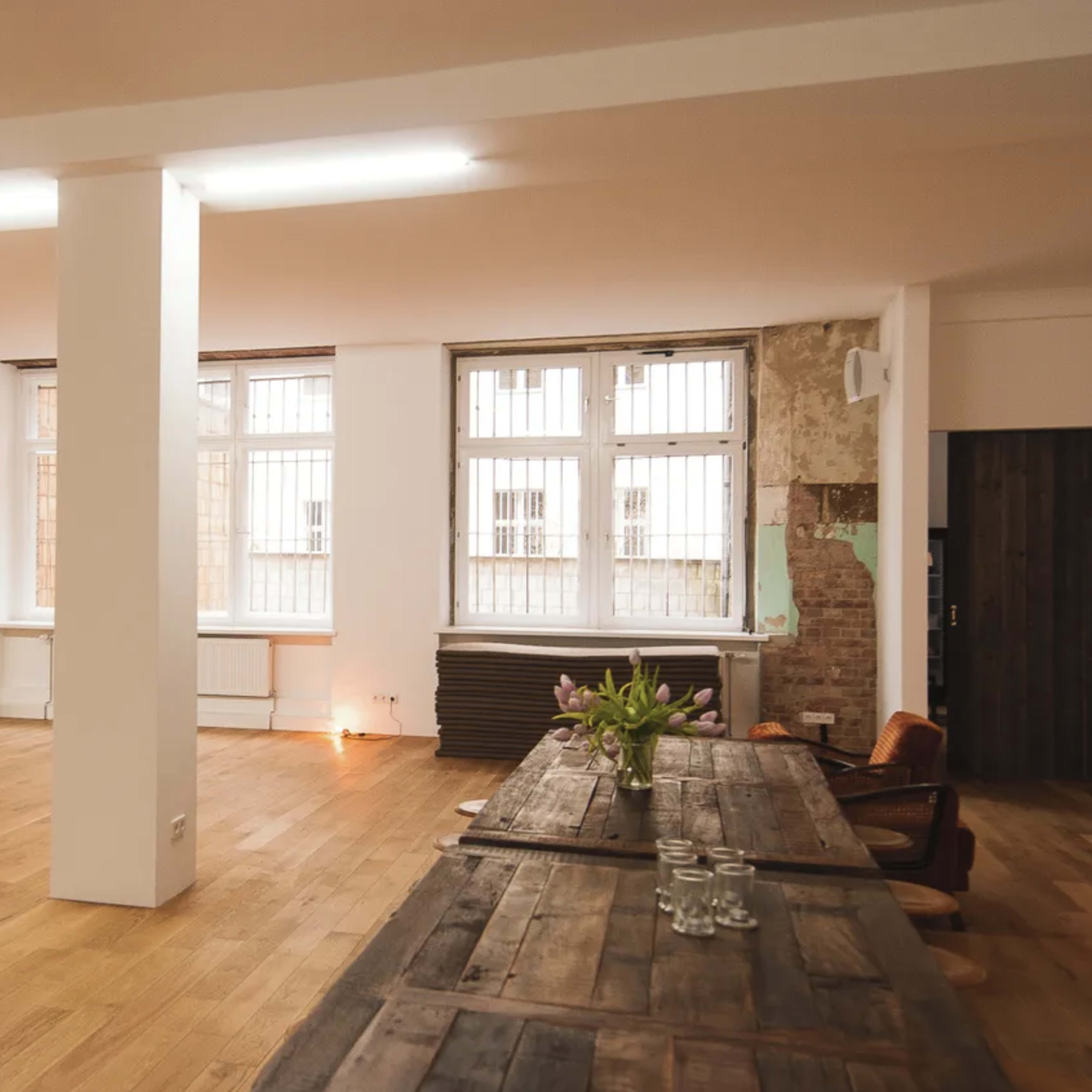 A spacious, minimalist room with wooden floors, large windows, and a long wooden table set with glasses.