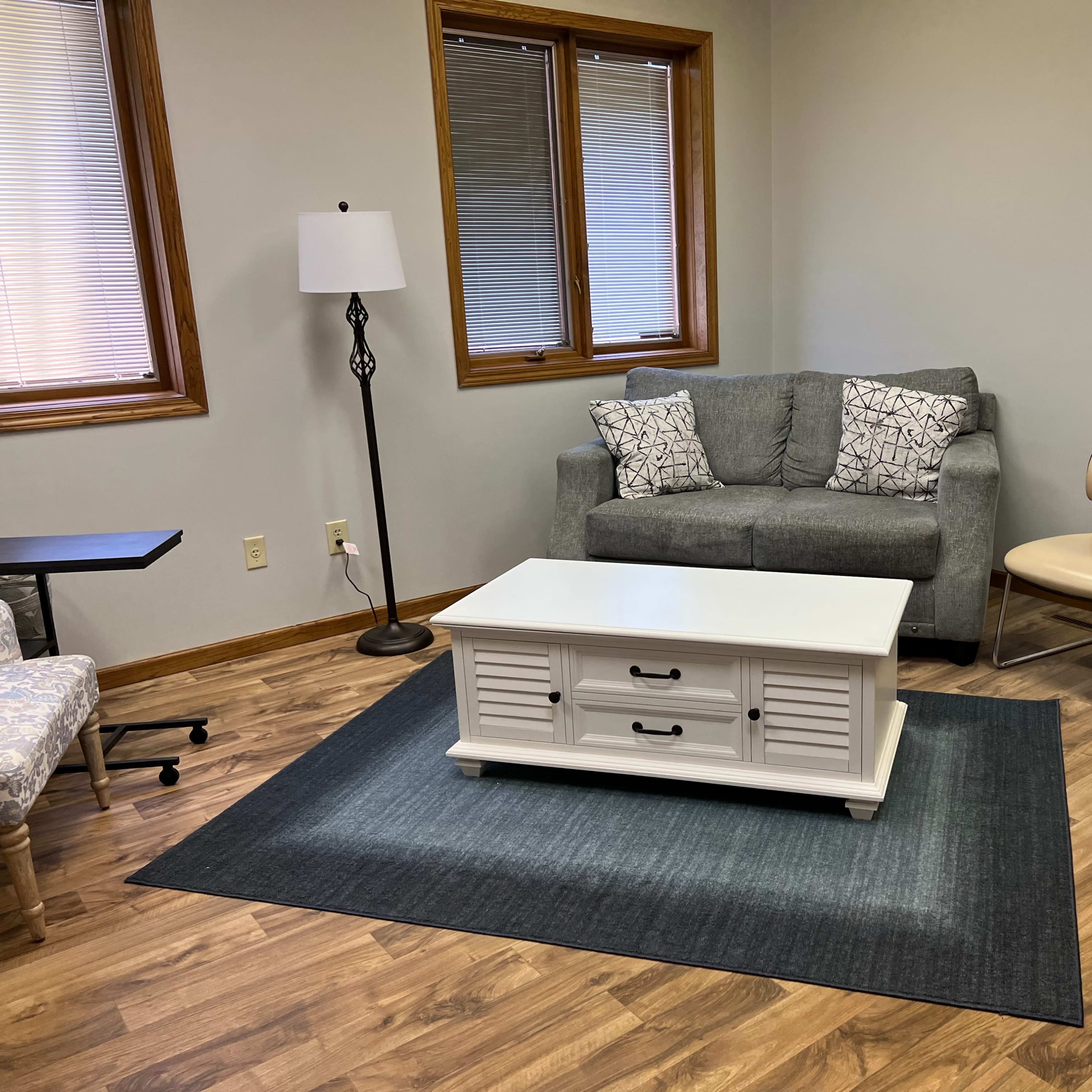 A small, furnished room with a gray sofa, a coffee table, a tan chair, and a lamp, all on a dark rug over a wooden floor.