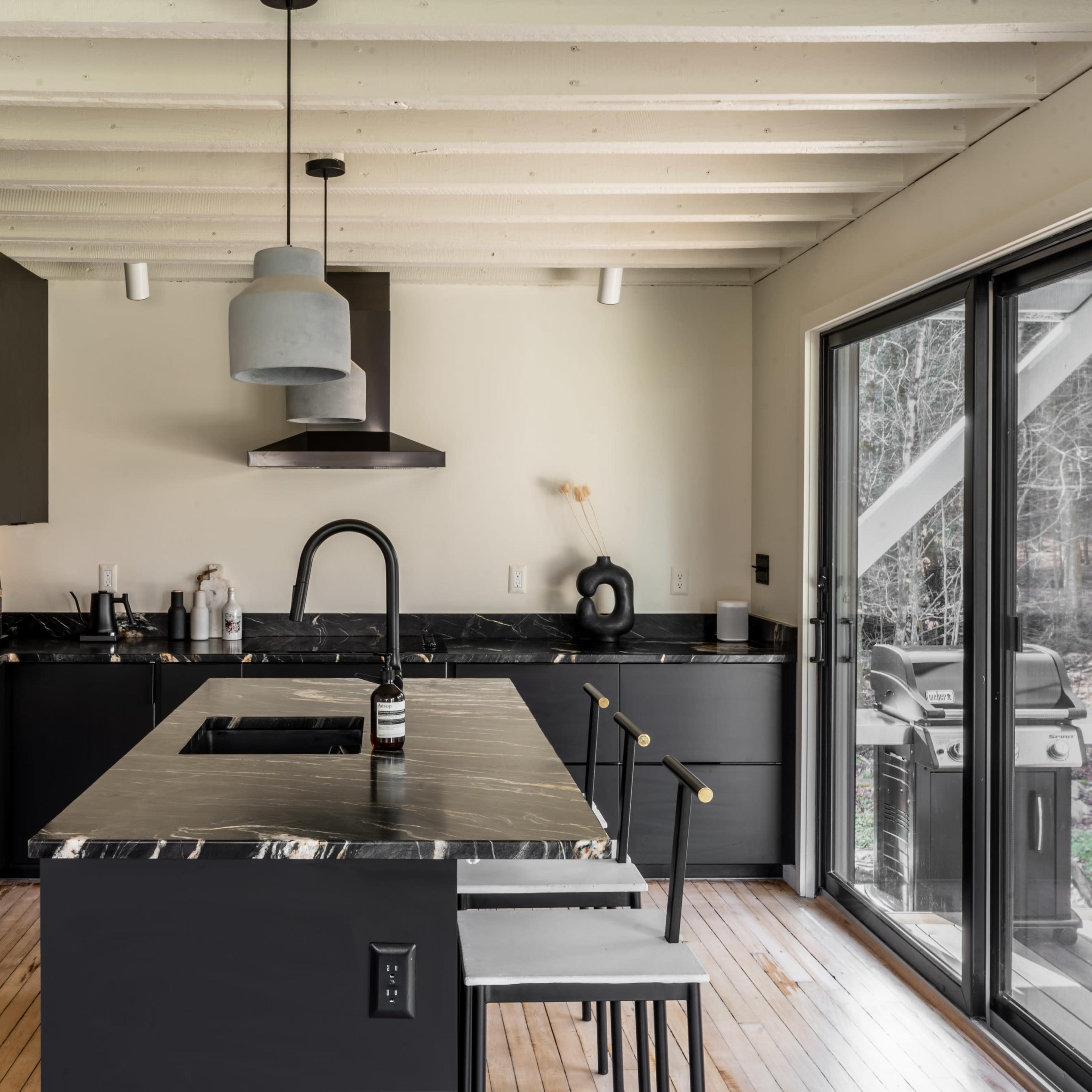 A modern kitchen features black cabinetry and a marble island with seating, overlooking a patio through sliding glass doors.
