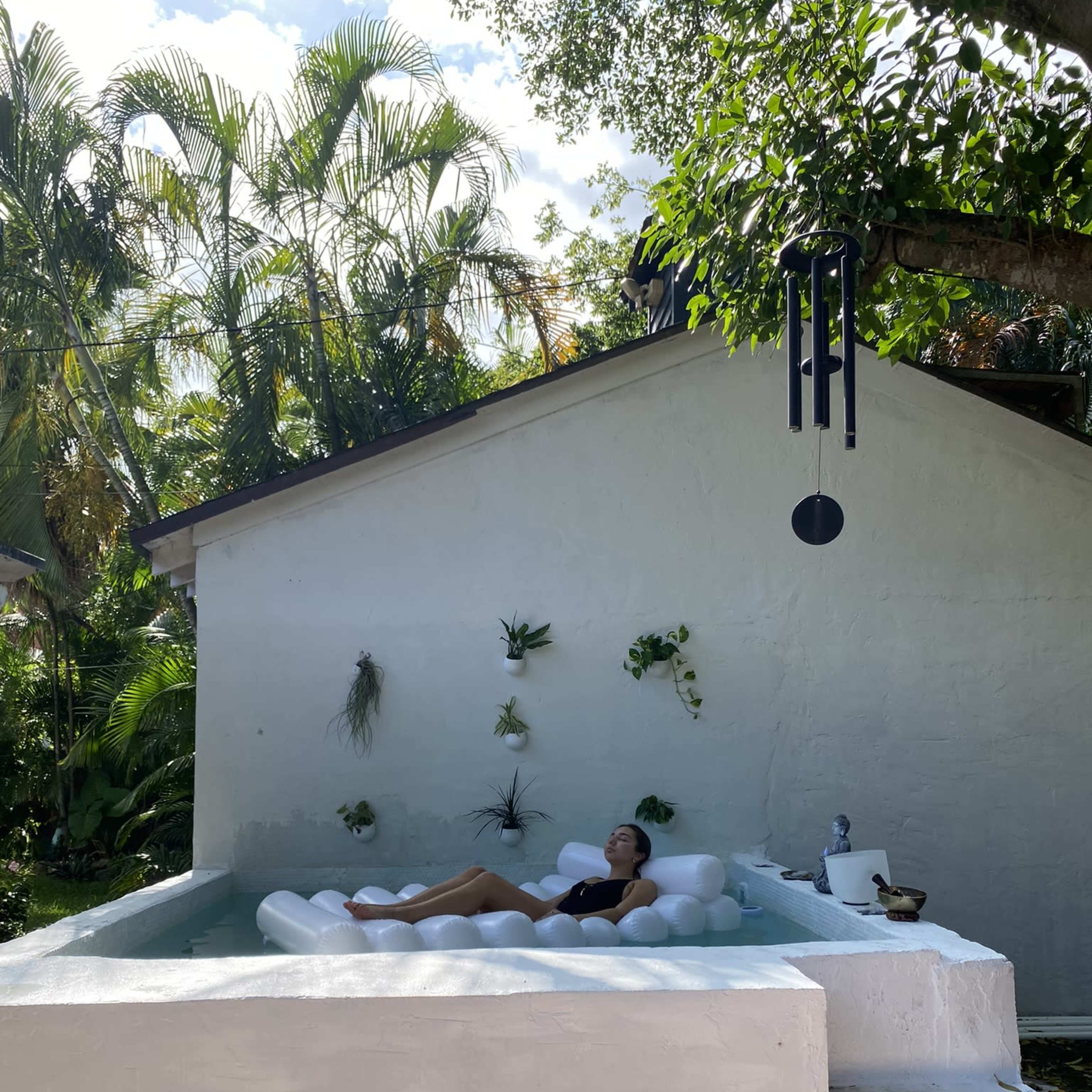 A person relaxes on a floating lounger in a small outdoor pool next to a white building surrounded by lush greenery.
