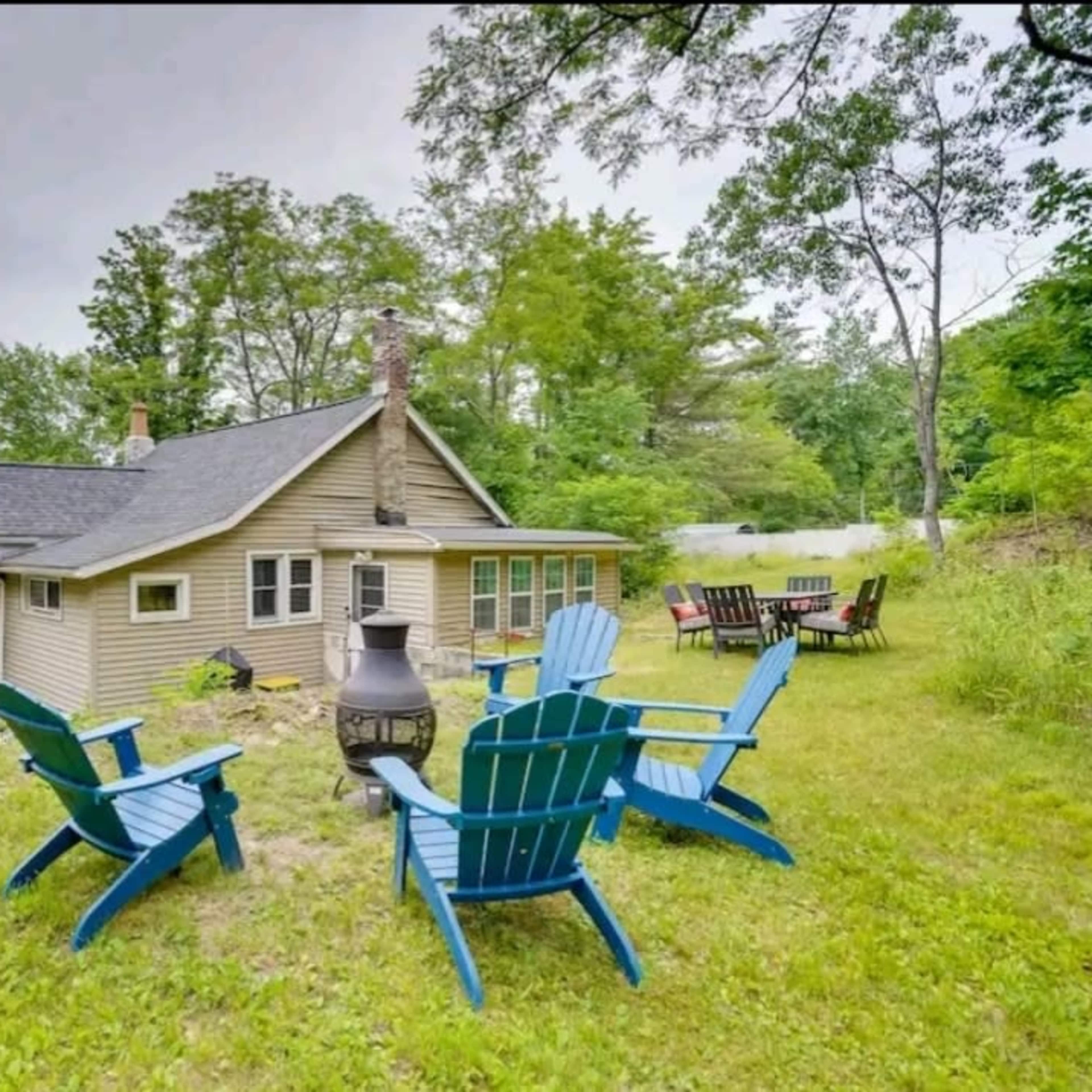 A beige house with a dark roof is surrounded by greenery, featuring several blue Adirondack chairs arranged around a fire pit in the backyard.