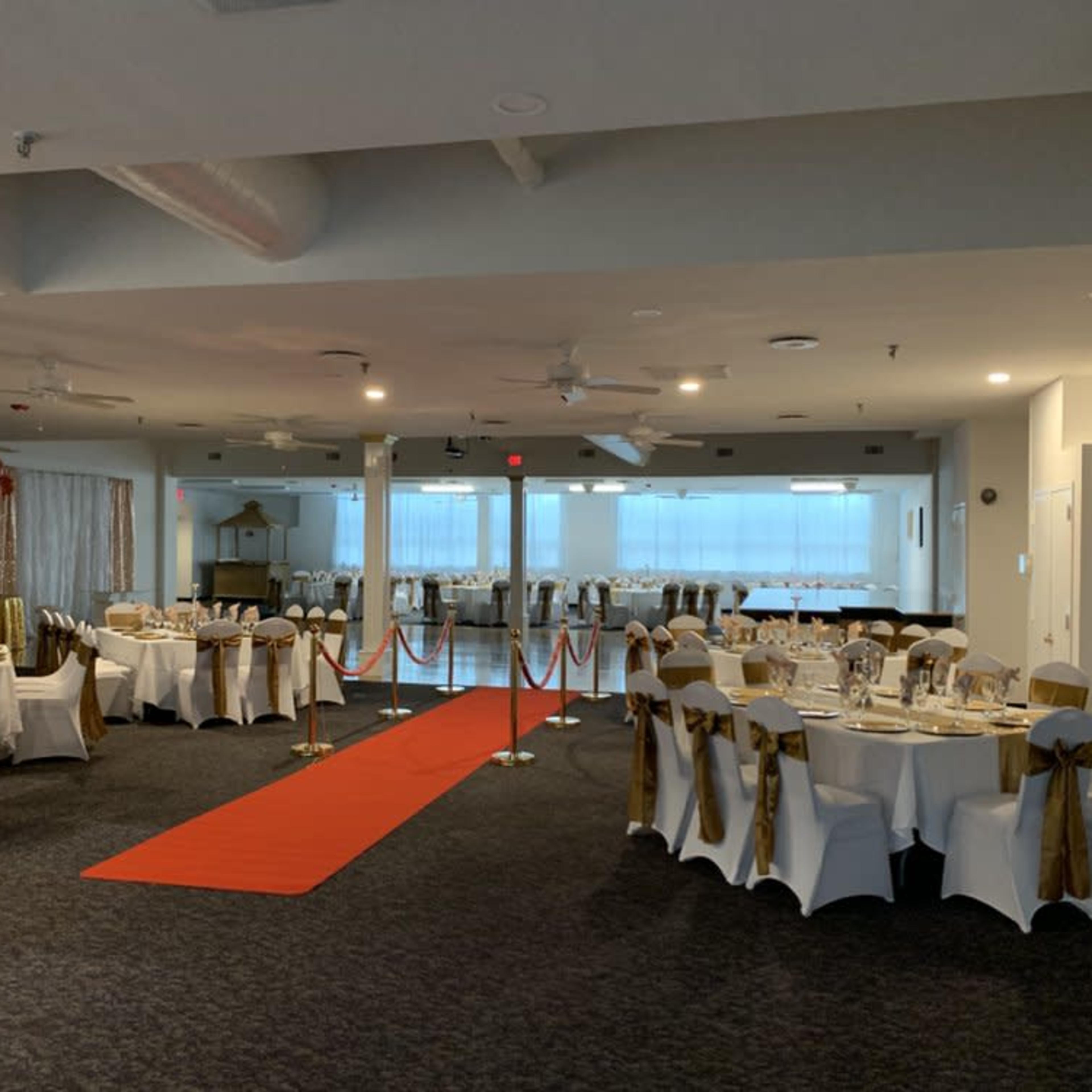 A banquet hall set up with tables draped in white and gold, featuring a red carpet leading to an area with large windows.