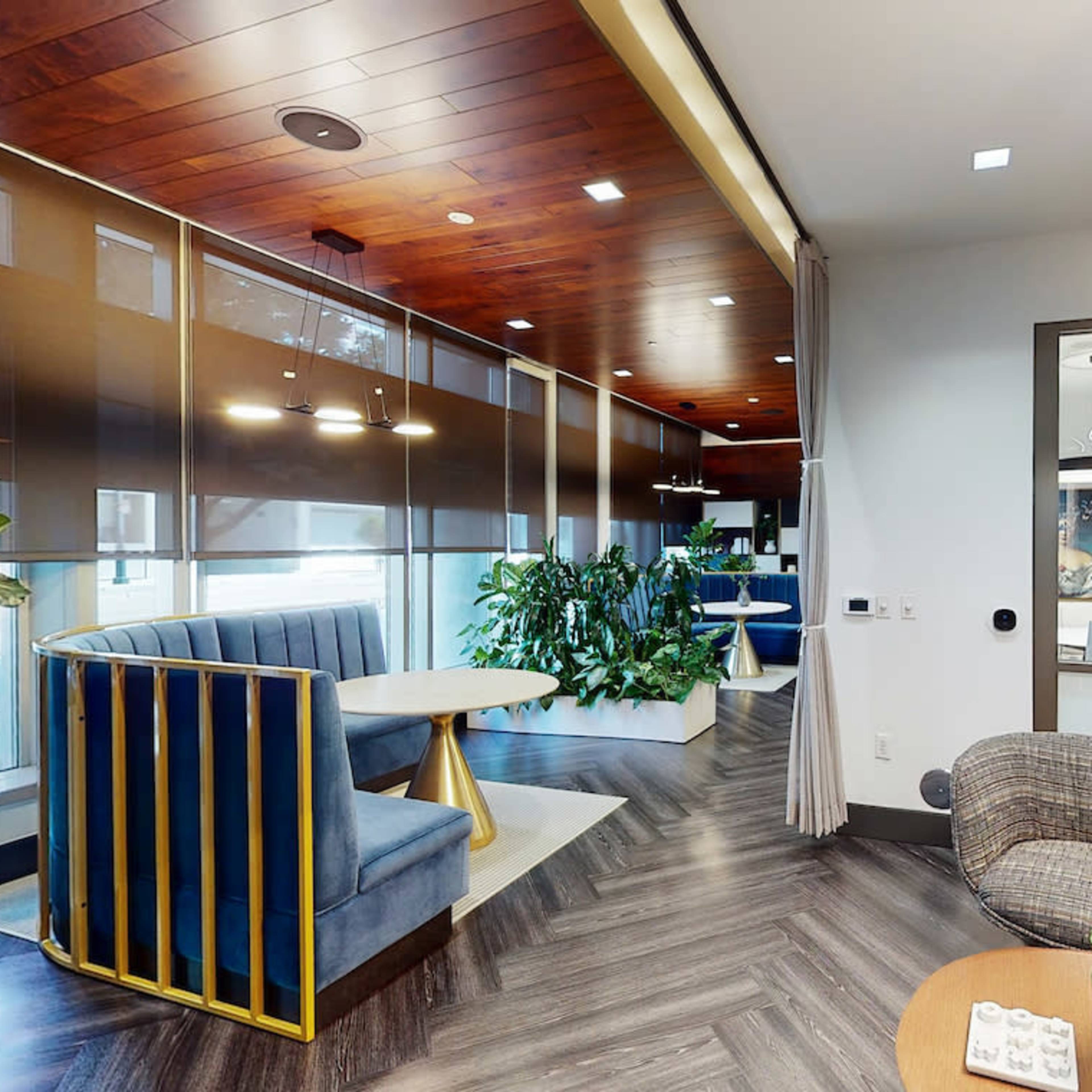 A modern office space featuring a lounge area with blue seating, a round table, and various indoor plants, all illuminated by ambient lighting.