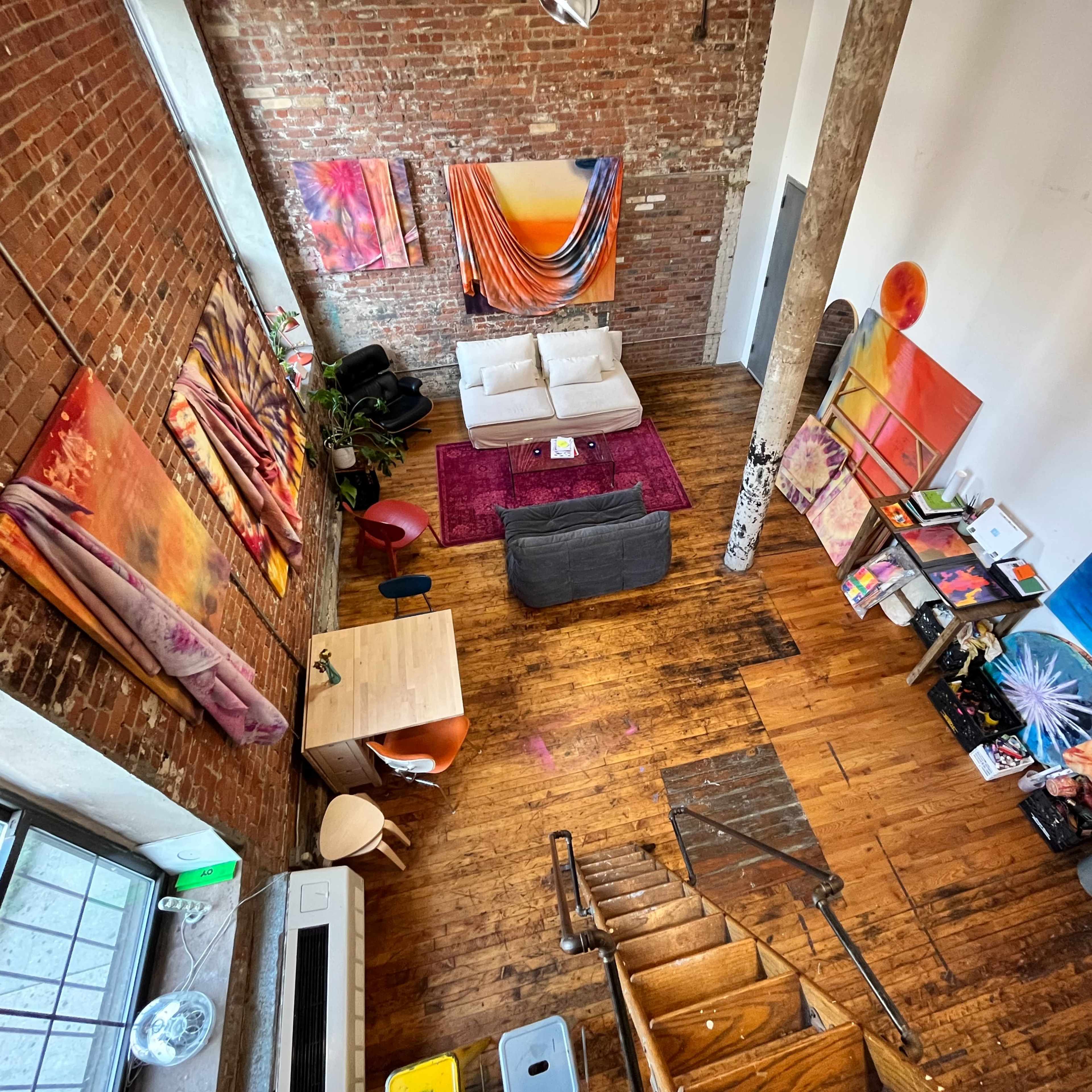 A spacious, loft-like living area featuring exposed brick walls, wooden flooring, and colorful artwork, with a seating arrangement and a staircase leading to an upper level.