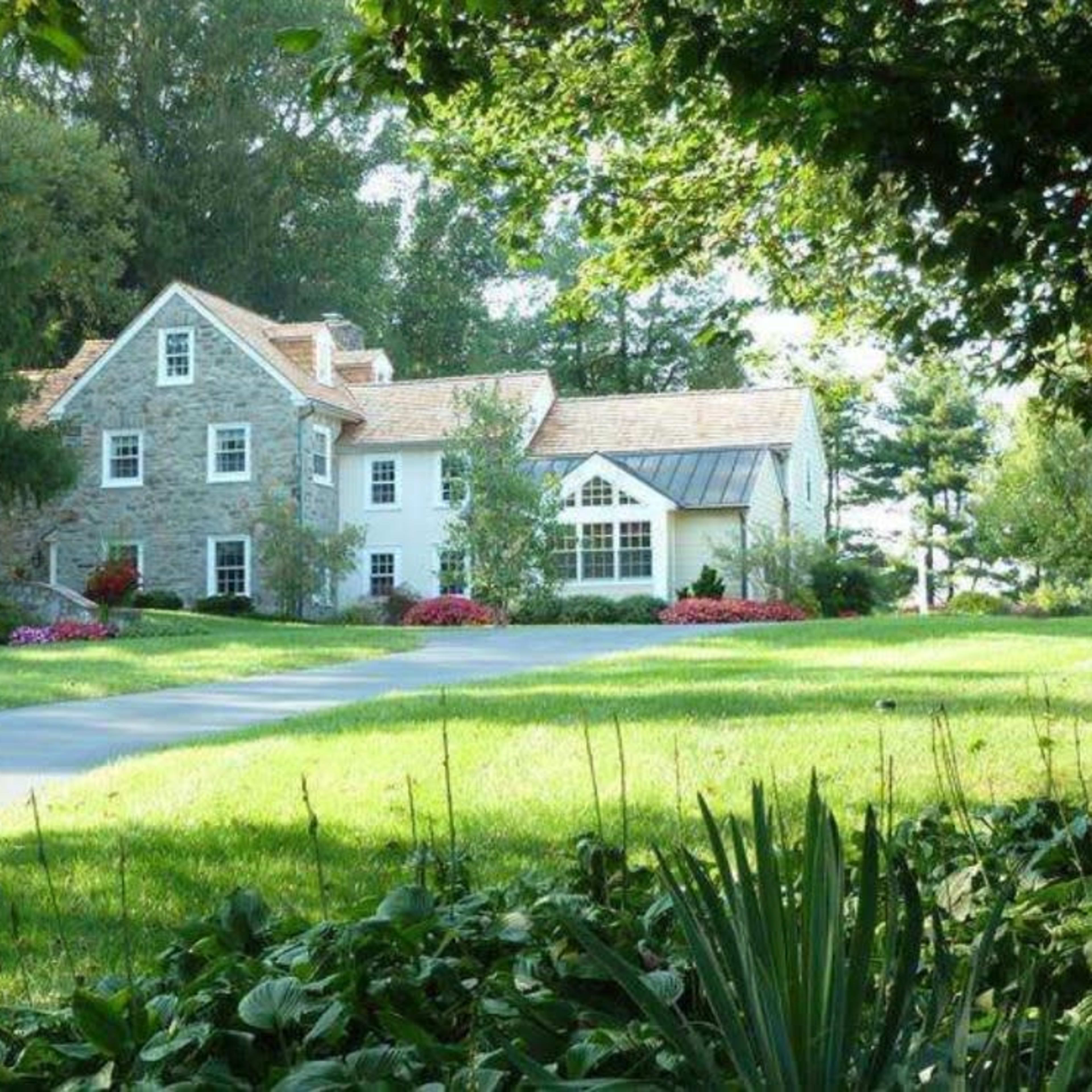 A stone house with a landscaped yard and a long driveway is surrounded by trees and flowers.
