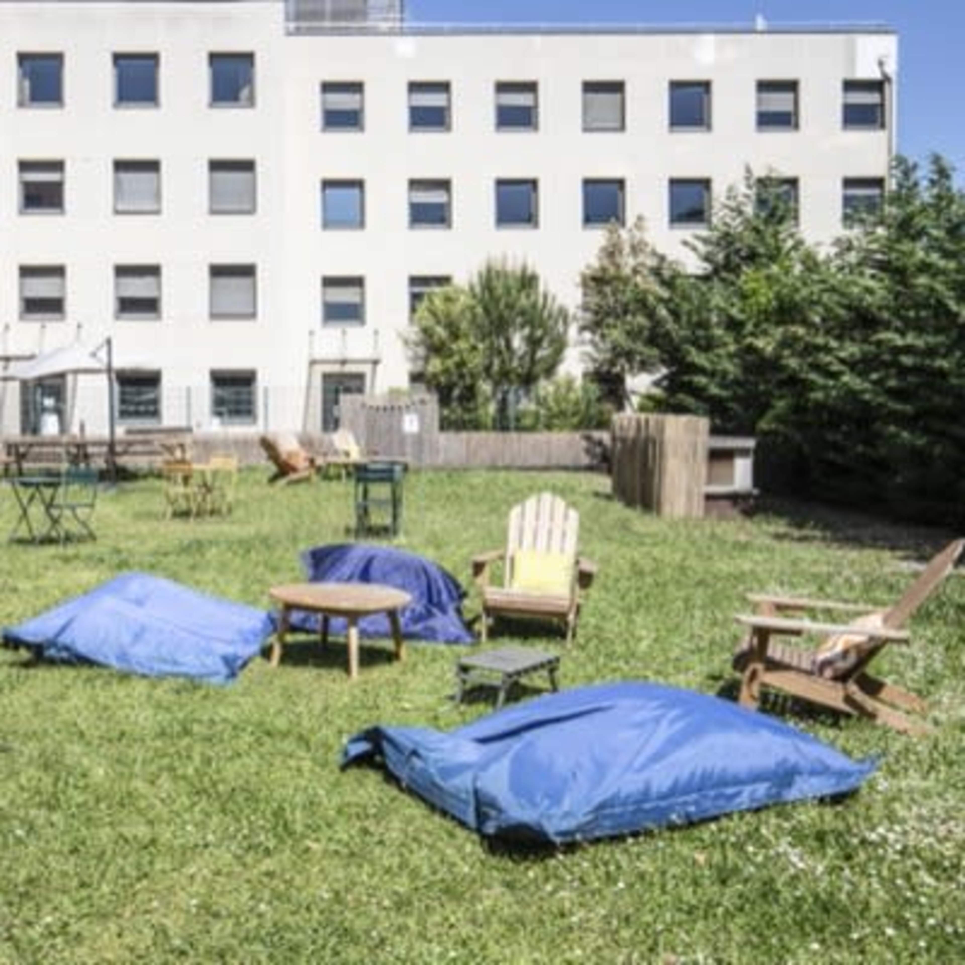 An outdoor area with blue cushions, wooden chairs, and tables on a grassy lawn surrounded by a building with multiple windows.