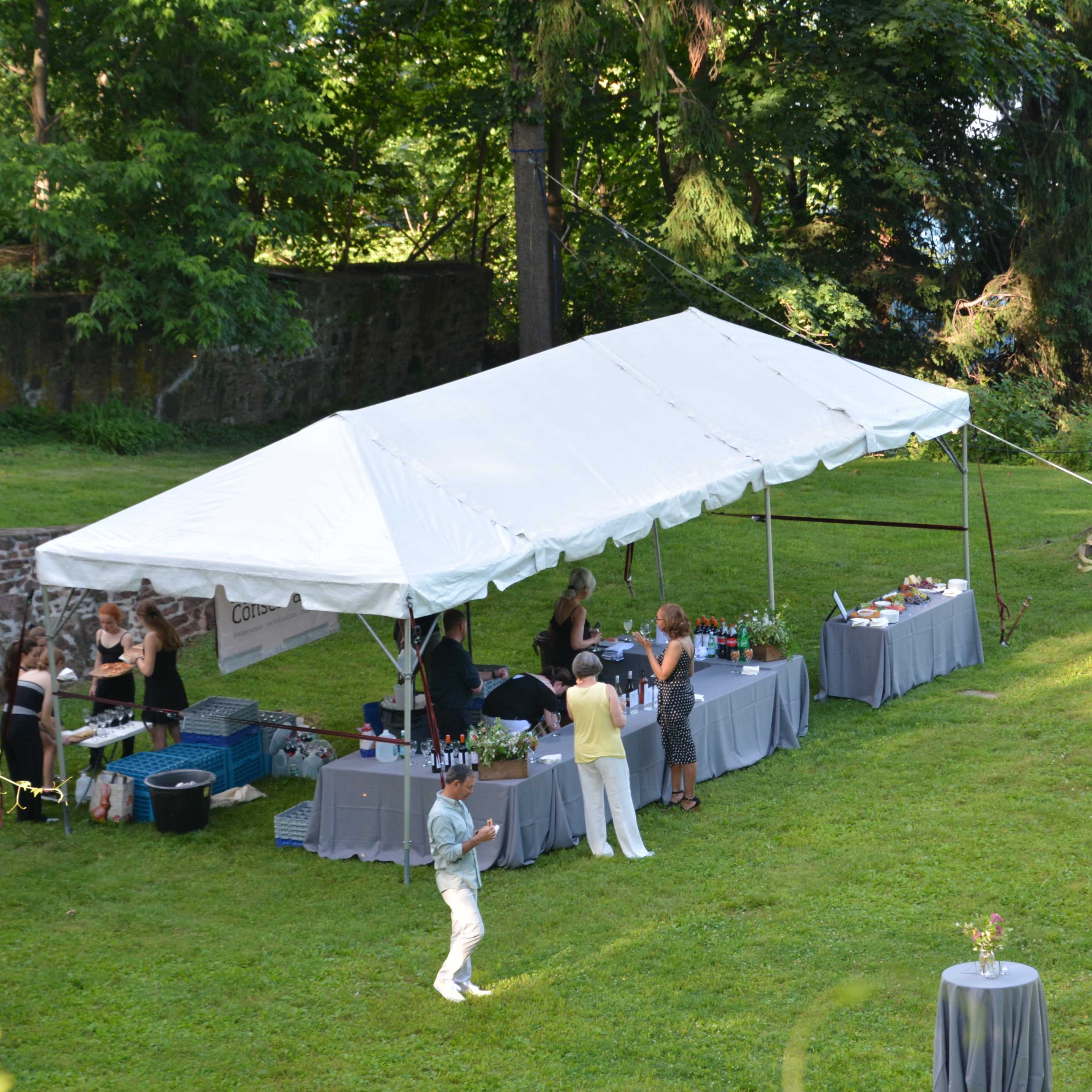 A tented outdoor event features several tables with drinks and food, surrounded by trees and a grassy area.