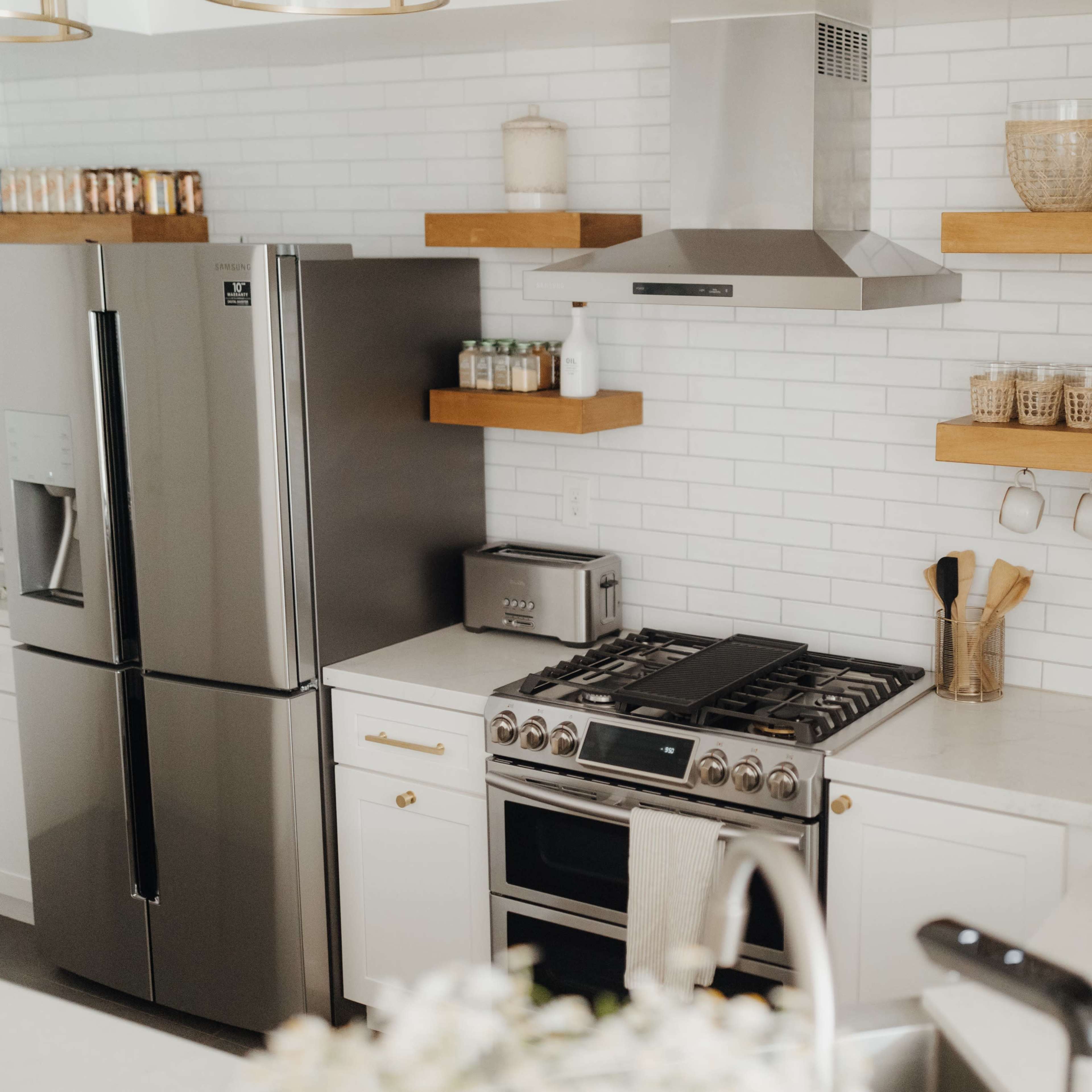A modern kitchen with a stainless steel refrigerator, a gas stove, and open shelves displaying jars and kitchen utensils against a white tiled wall.