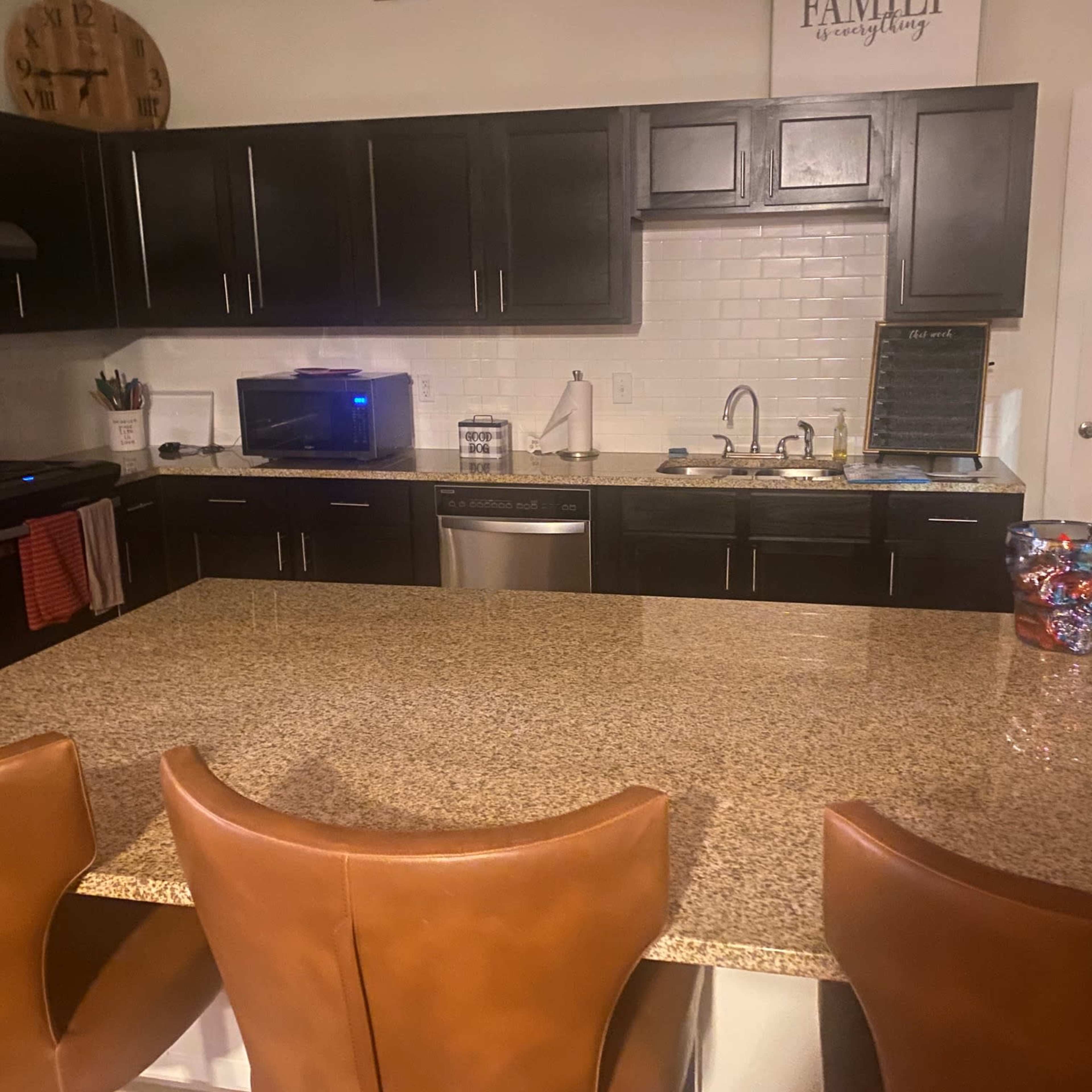 A modern kitchen features dark cabinets, a large granite island with four brown leather stools, and stainless steel appliances.