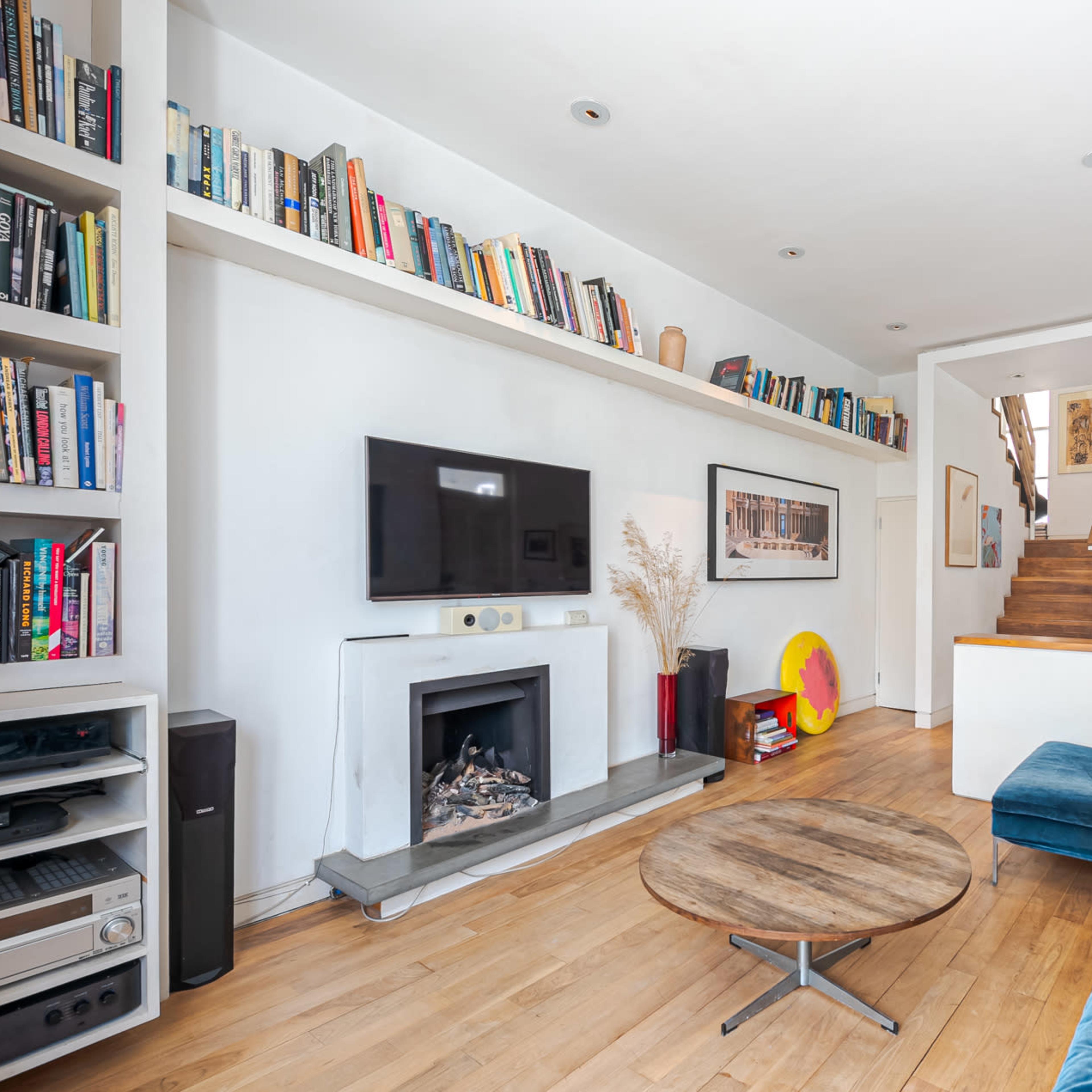 A living room features a blue sectional sofa, a wooden coffee table, a television mounted on the wall, and bookshelves filled with books along the upper wall.