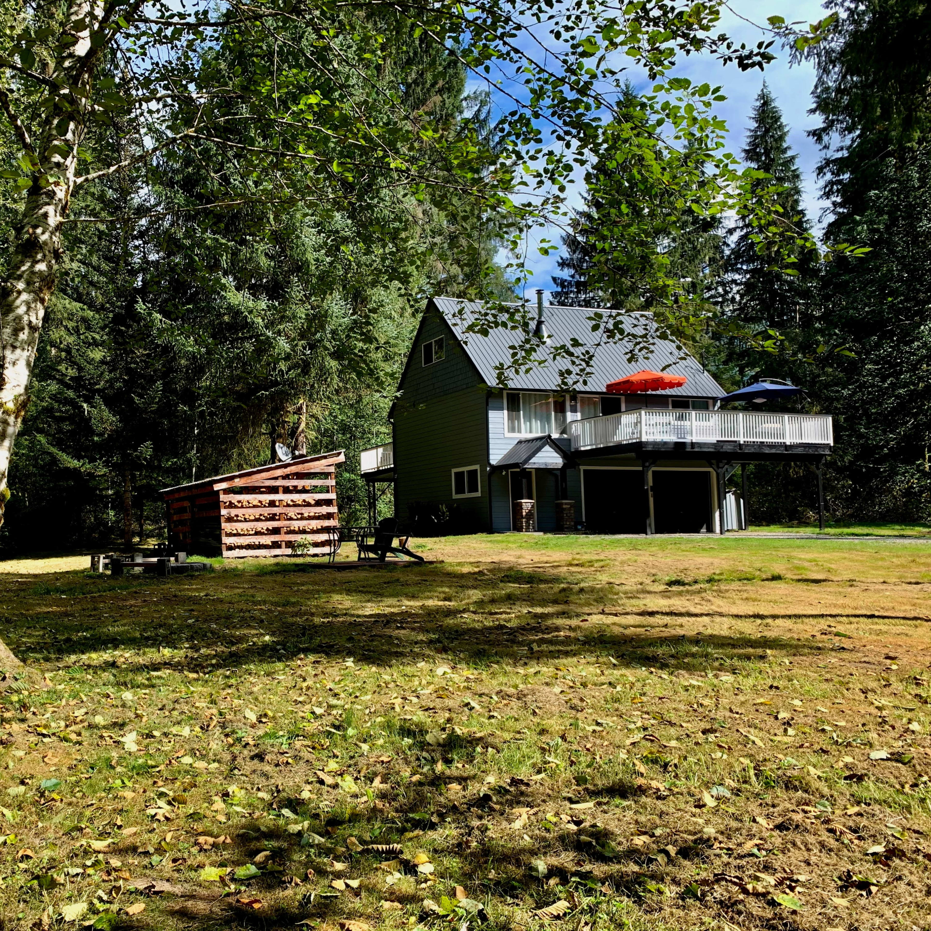 A green two-story house with a deck and an umbrella sits surrounded by trees in a grassy clearing.