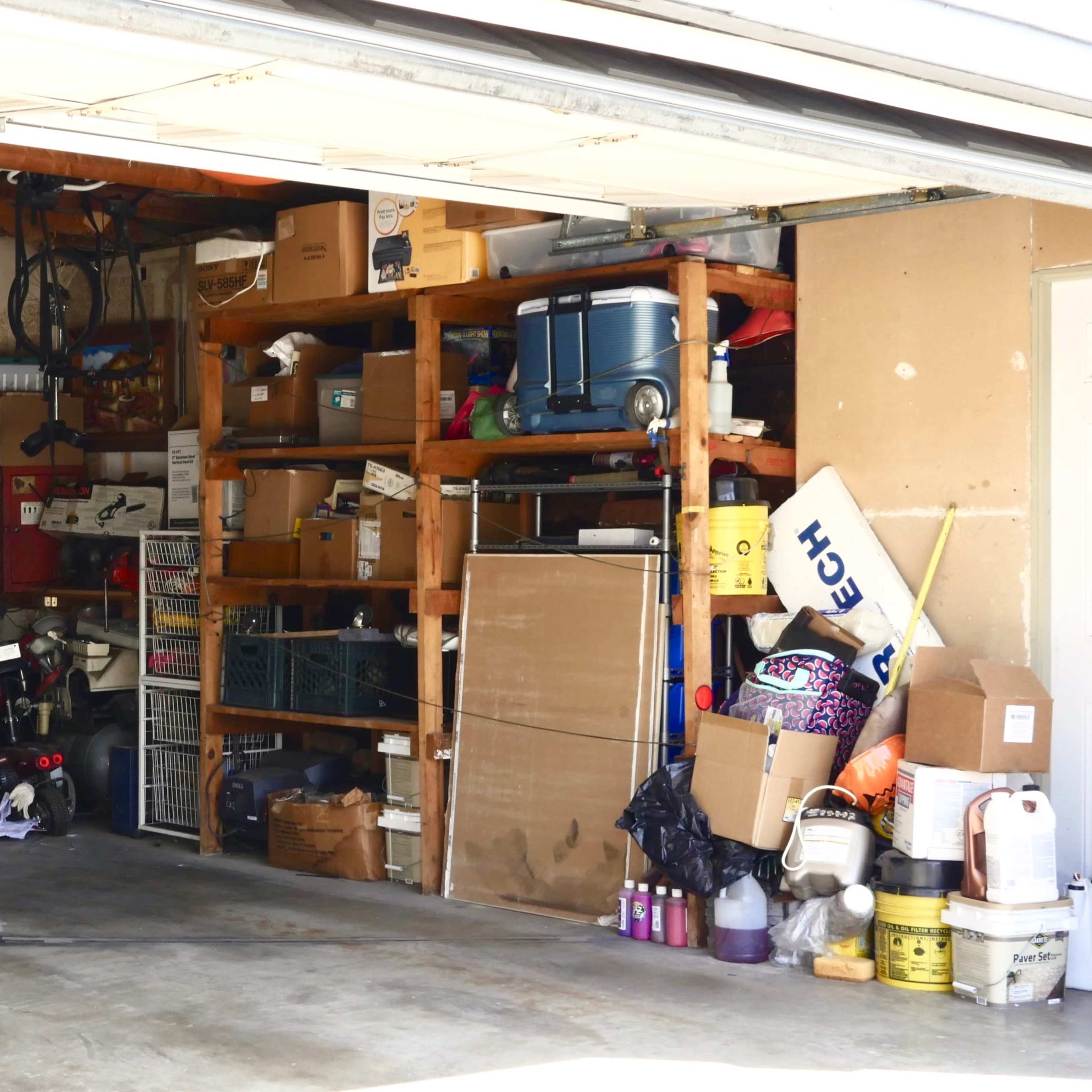 A garage is filled with shelves of boxes, tools, and various storage items, with a refrigerator in the corner and an assortment of clutter on the floor.