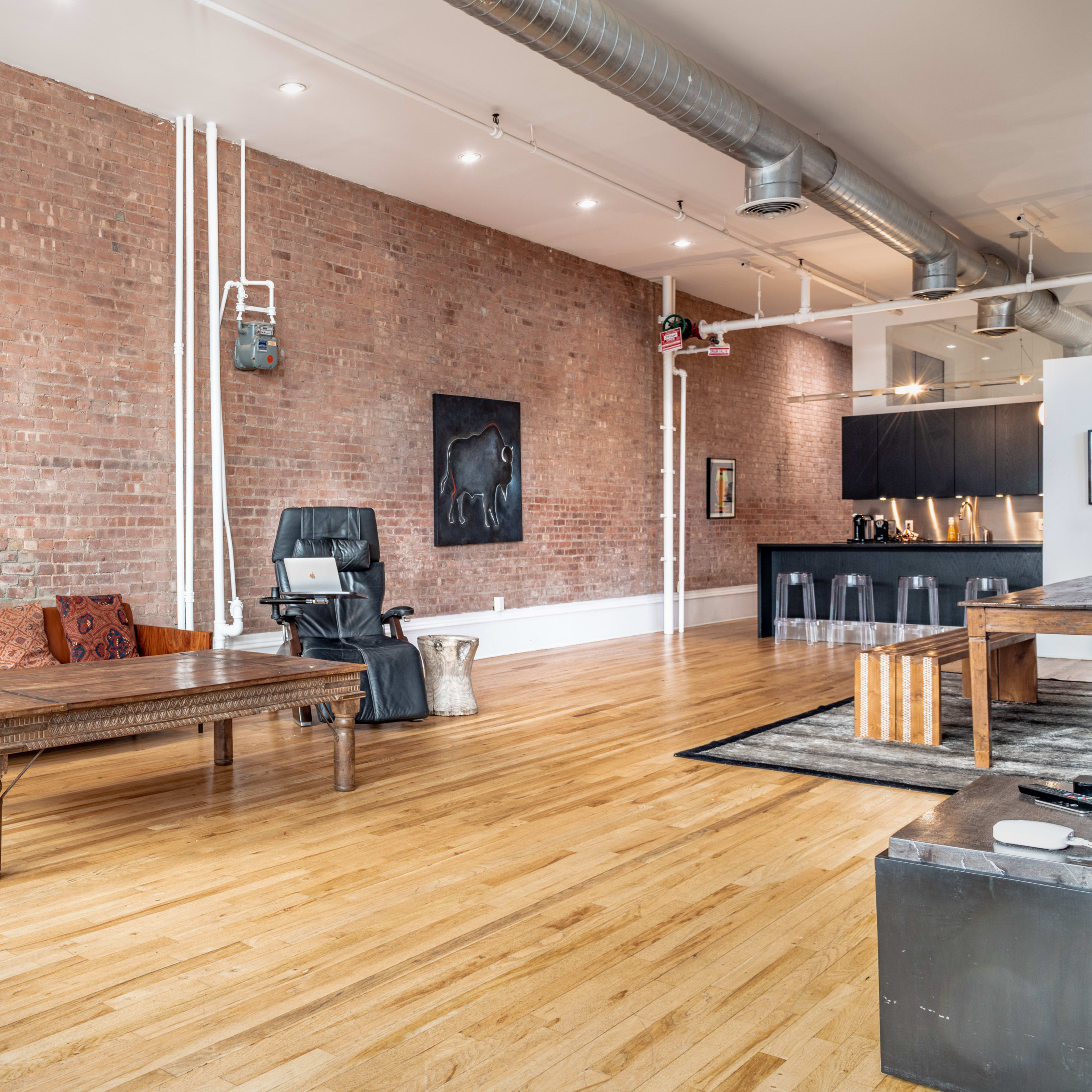 A spacious living area with exposed brick walls, wooden flooring, a brown sofa, a large coffee table, and a kitchen area in the background.