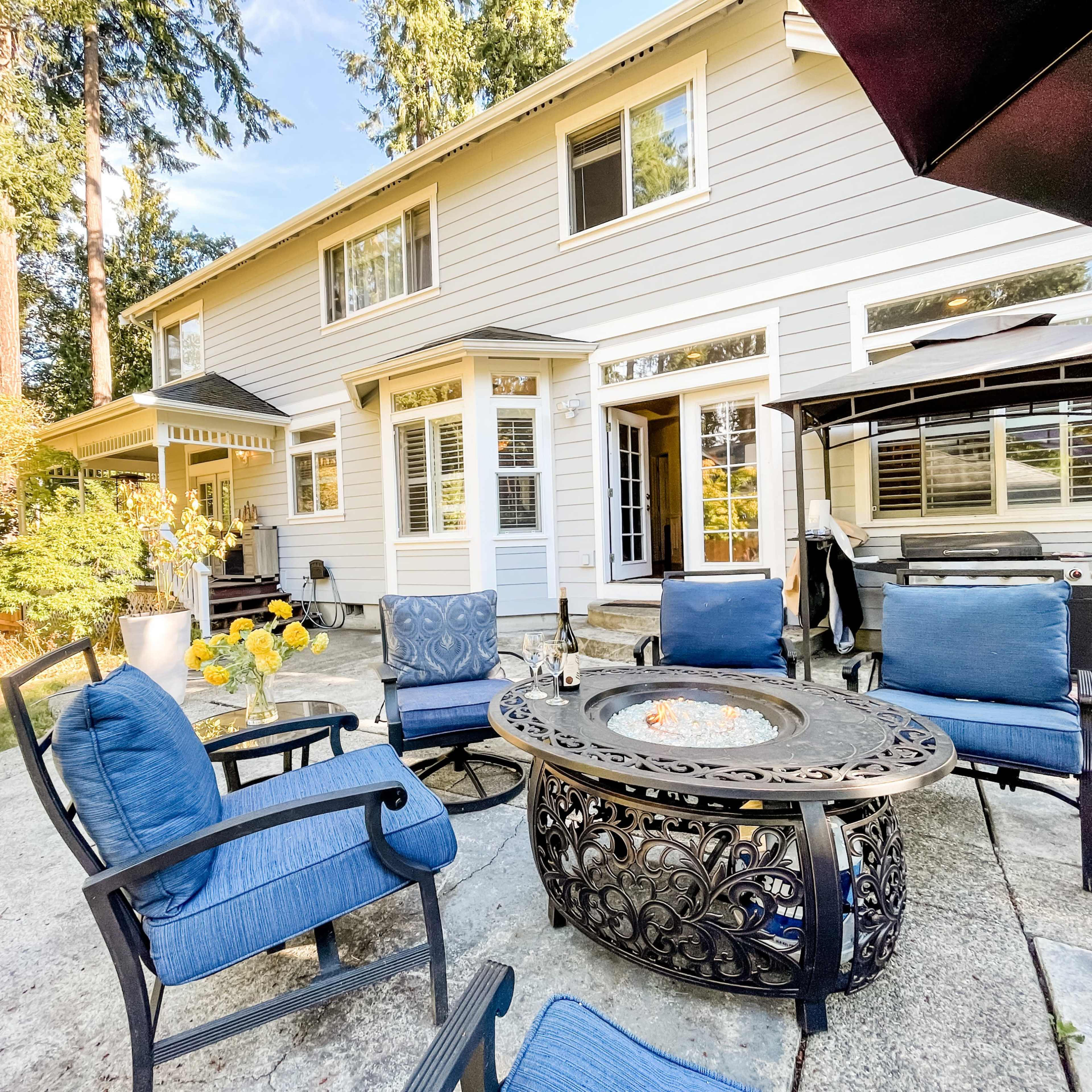 A backyard patio area featuring a fire pit surrounded by blue chairs and a house with large windows in the background.