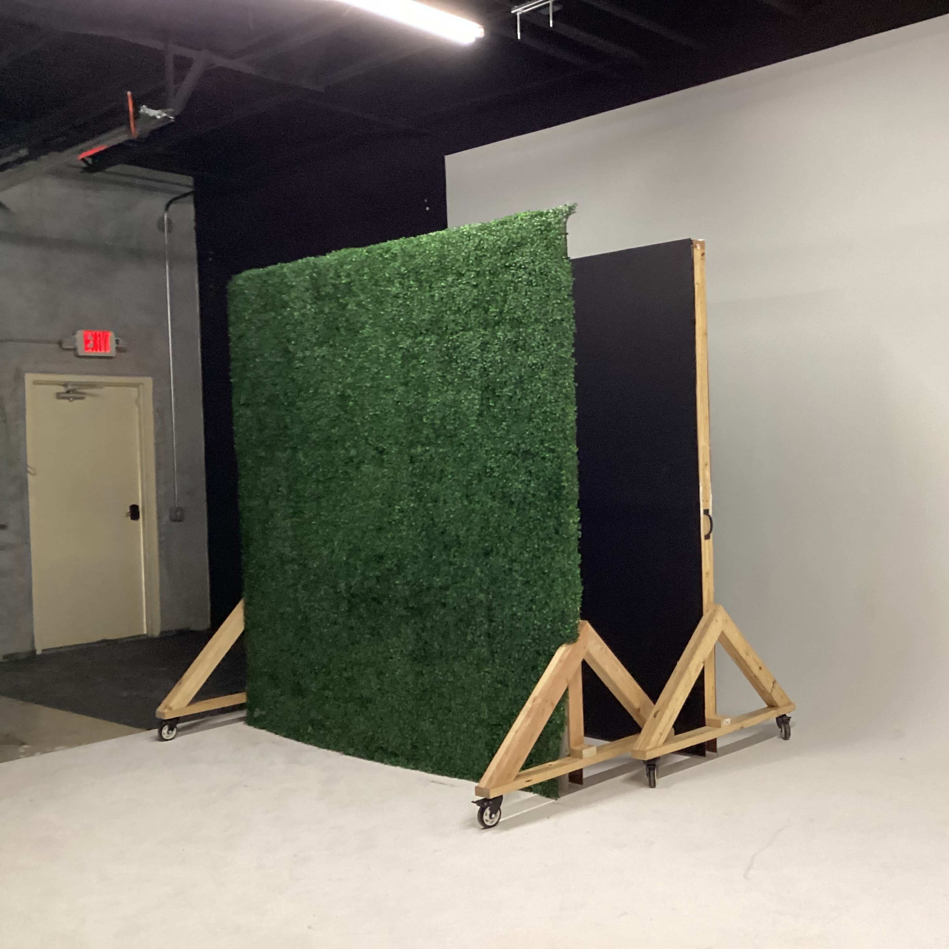 A green artificial grass wall is positioned on a wooden frame with wheels against a plain gray backdrop in a studio space.