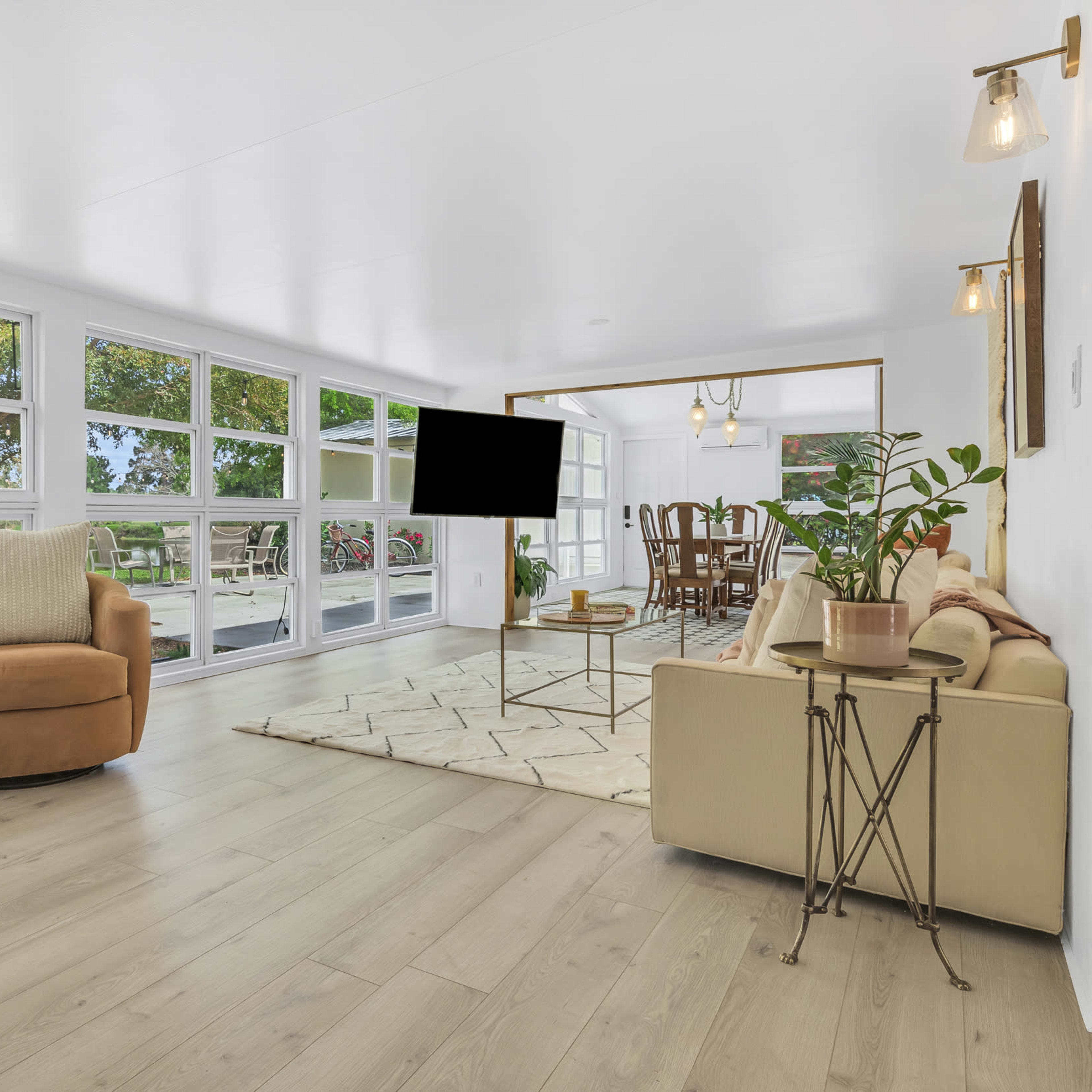 A bright and spacious living area features large windows overlooking a view, with a cozy armchair, a sofa, and a TV mounted on the wall.