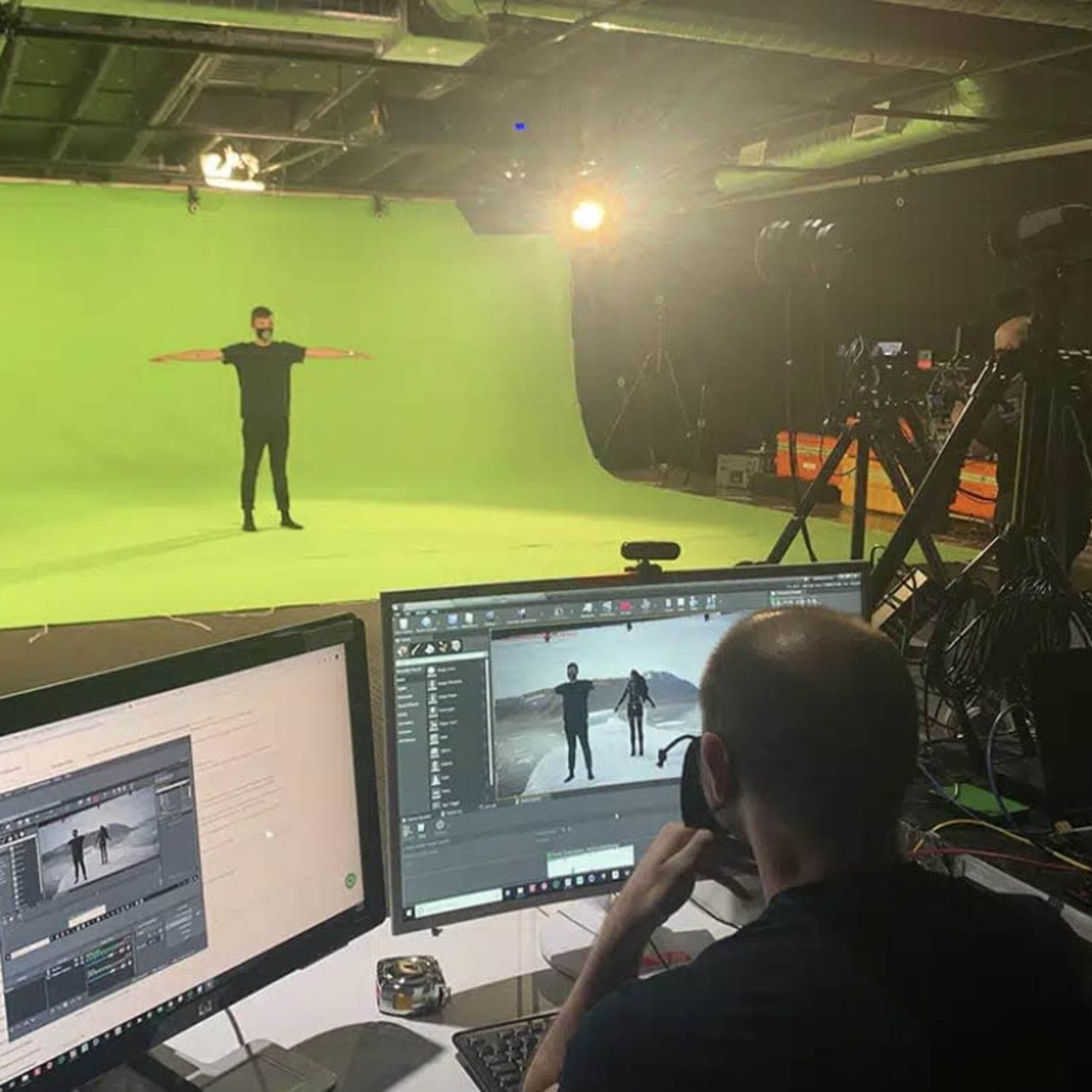 A person stands with arms outstretched in front of a green screen while another person works on a computer at a desk in a studio setting.