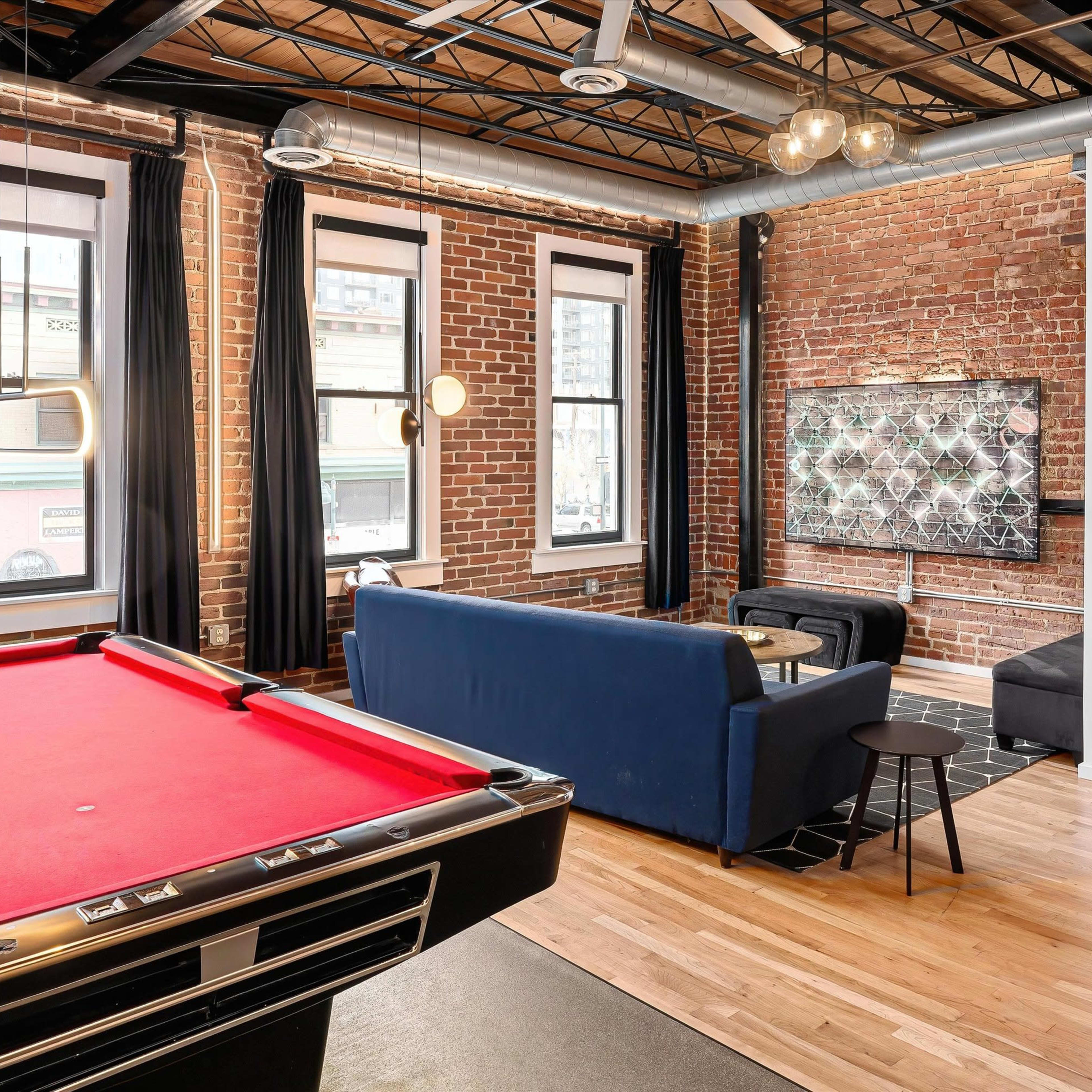A living space with a red pool table in the foreground, a blue sofa and a coffee table in the center, and large windows with black curtains along the brick wall.