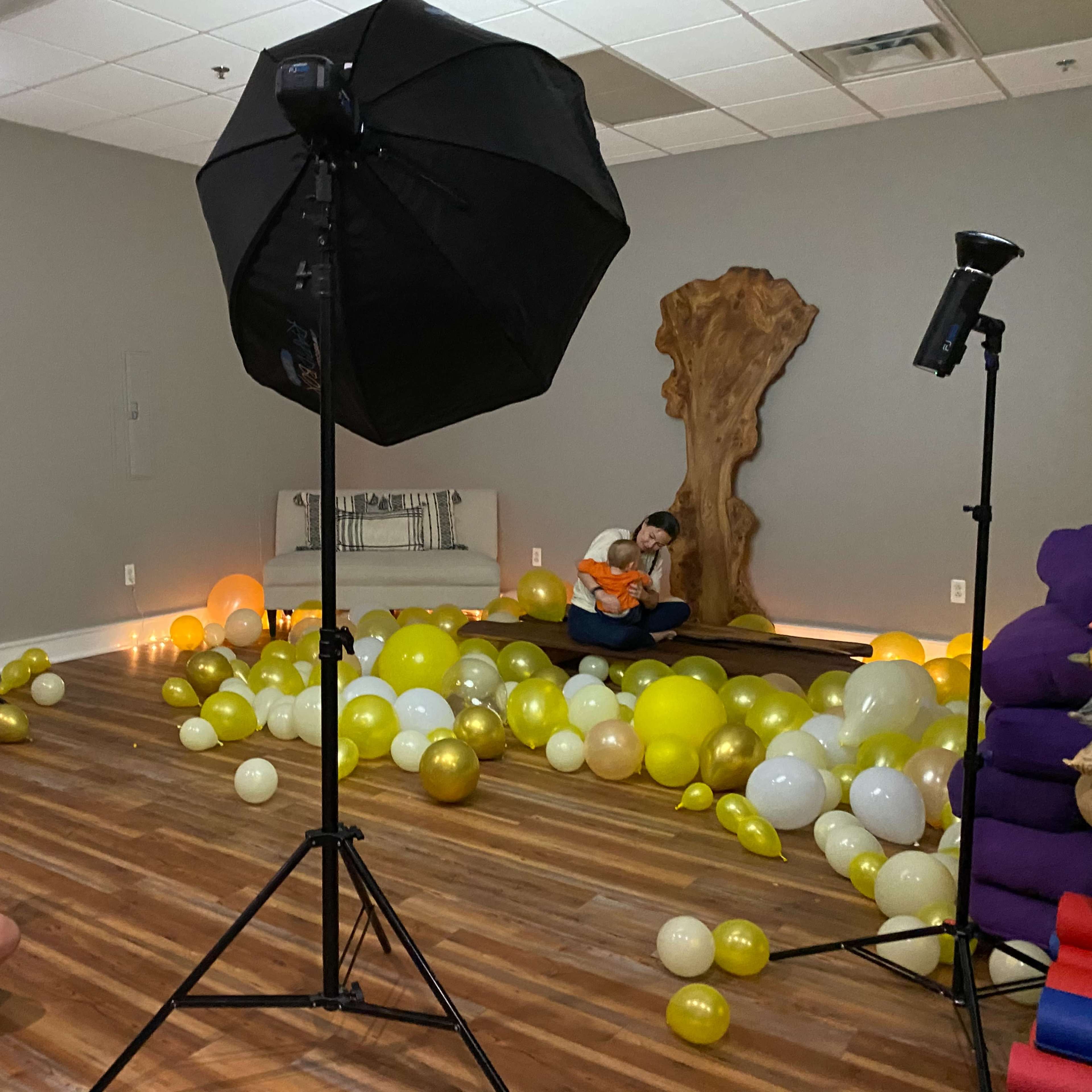 A photographer captures a child sitting amidst yellow and transparent balloons in a studio setting with softbox lighting.