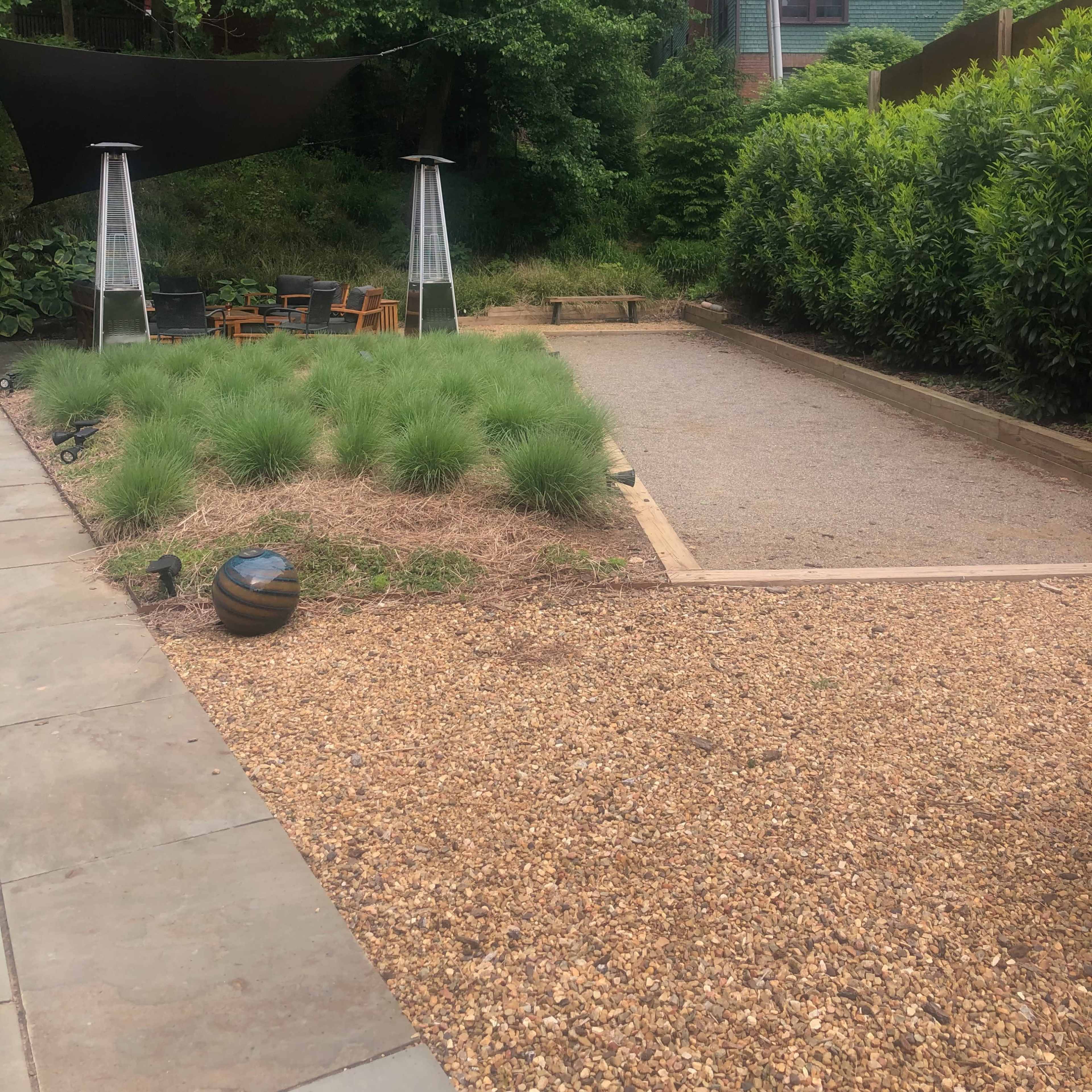 A landscaped outdoor area featuring a gravel path, low greenery, and two tall patio heaters under a shaded seating arrangement.