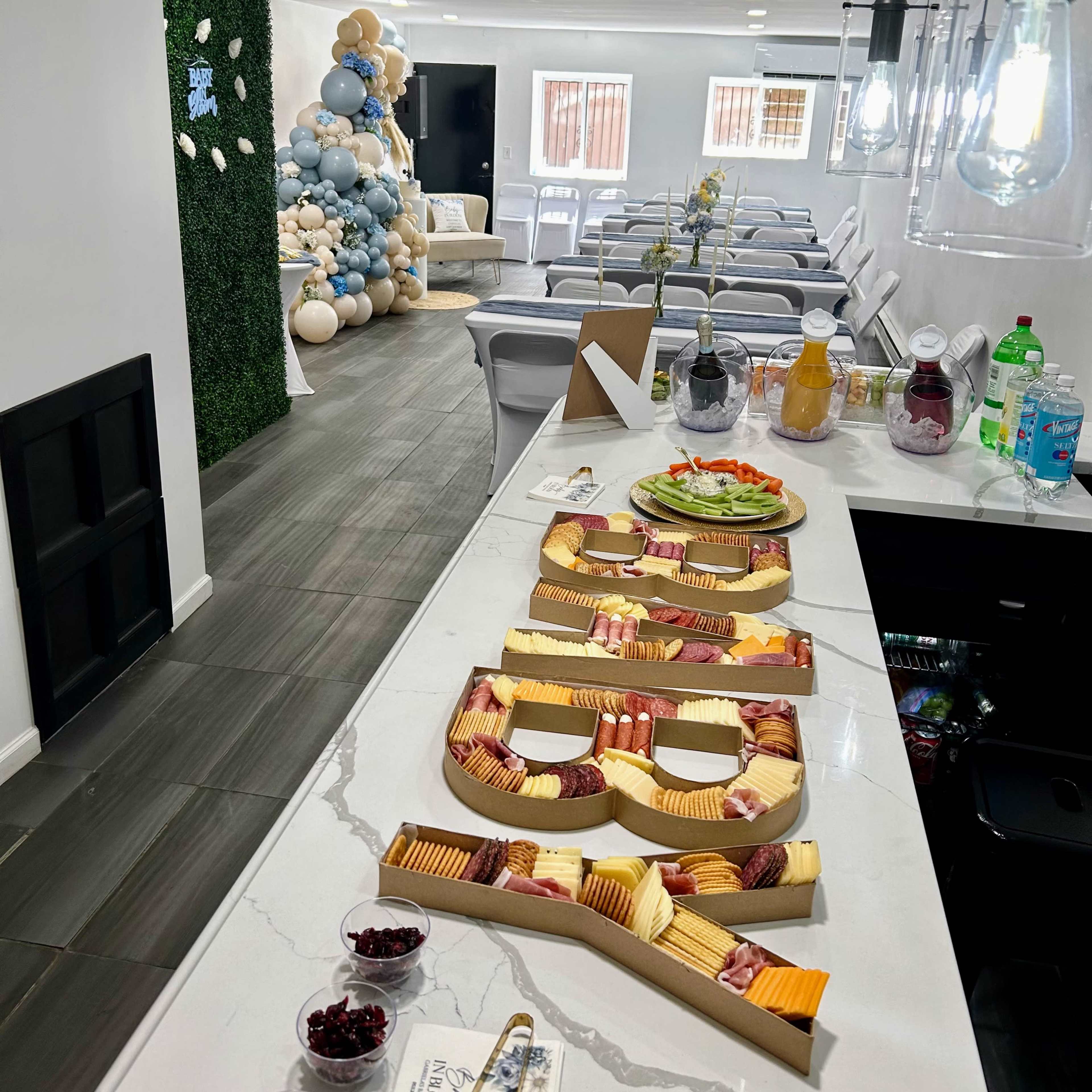 A long table displays a charcuterie board with various cheeses and meats, while a seating area and a balloon display are visible in the background.