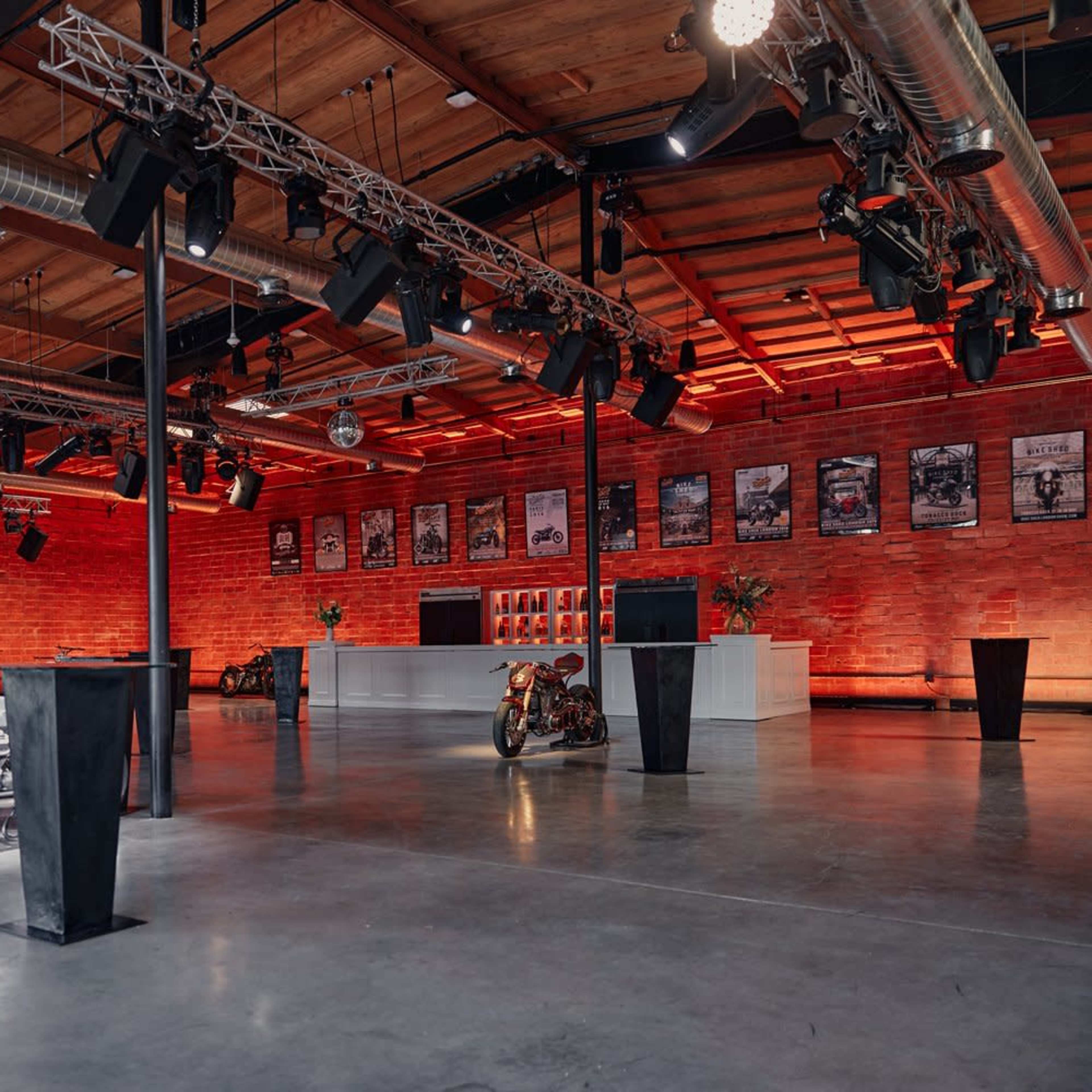 The spacious industrial venue features vintage motorcycles on display and walls adorned with posters, illuminated by red lighting.