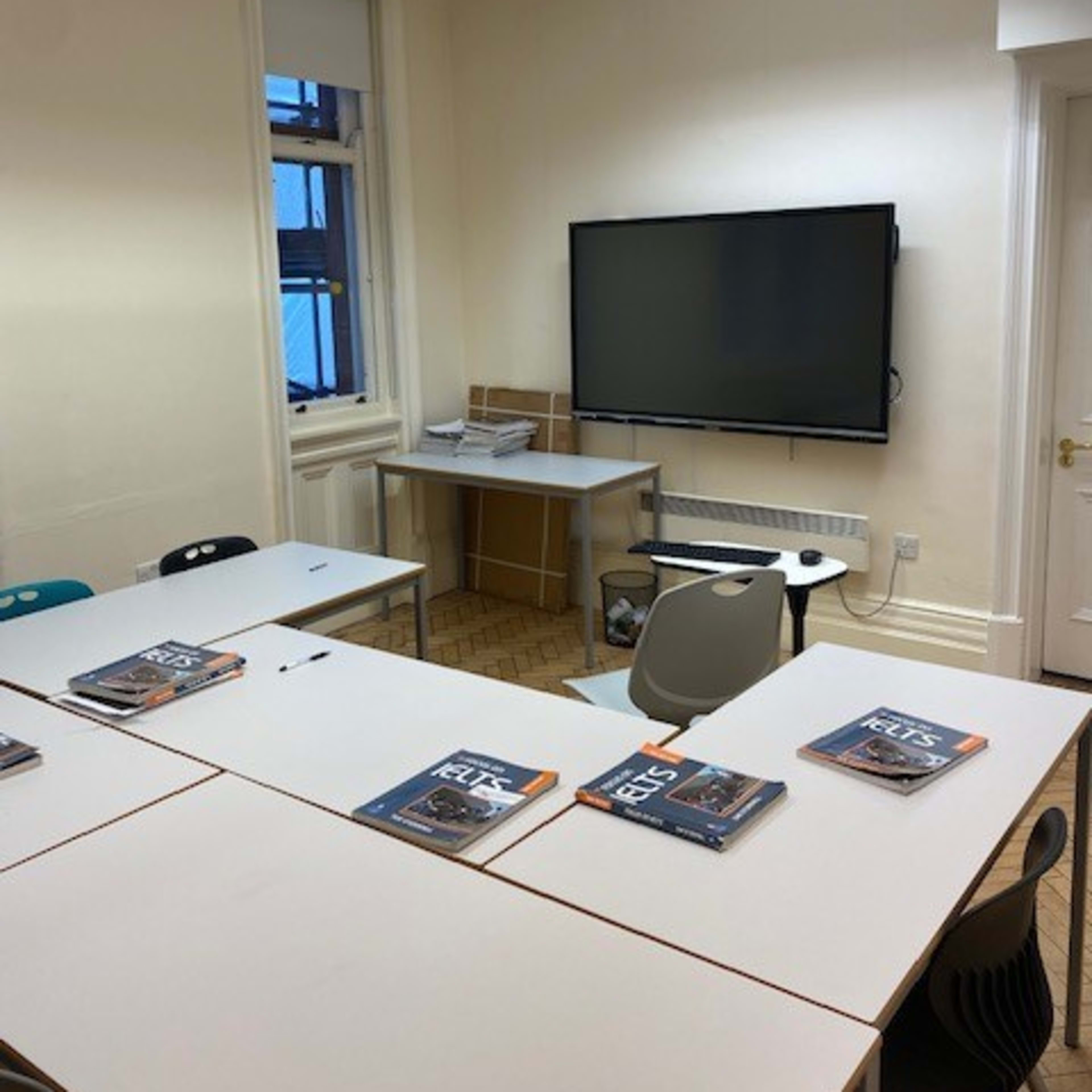 A classroom is arranged with several tables and chairs, along with a TV mounted on the wall and textbooks placed on the tables.