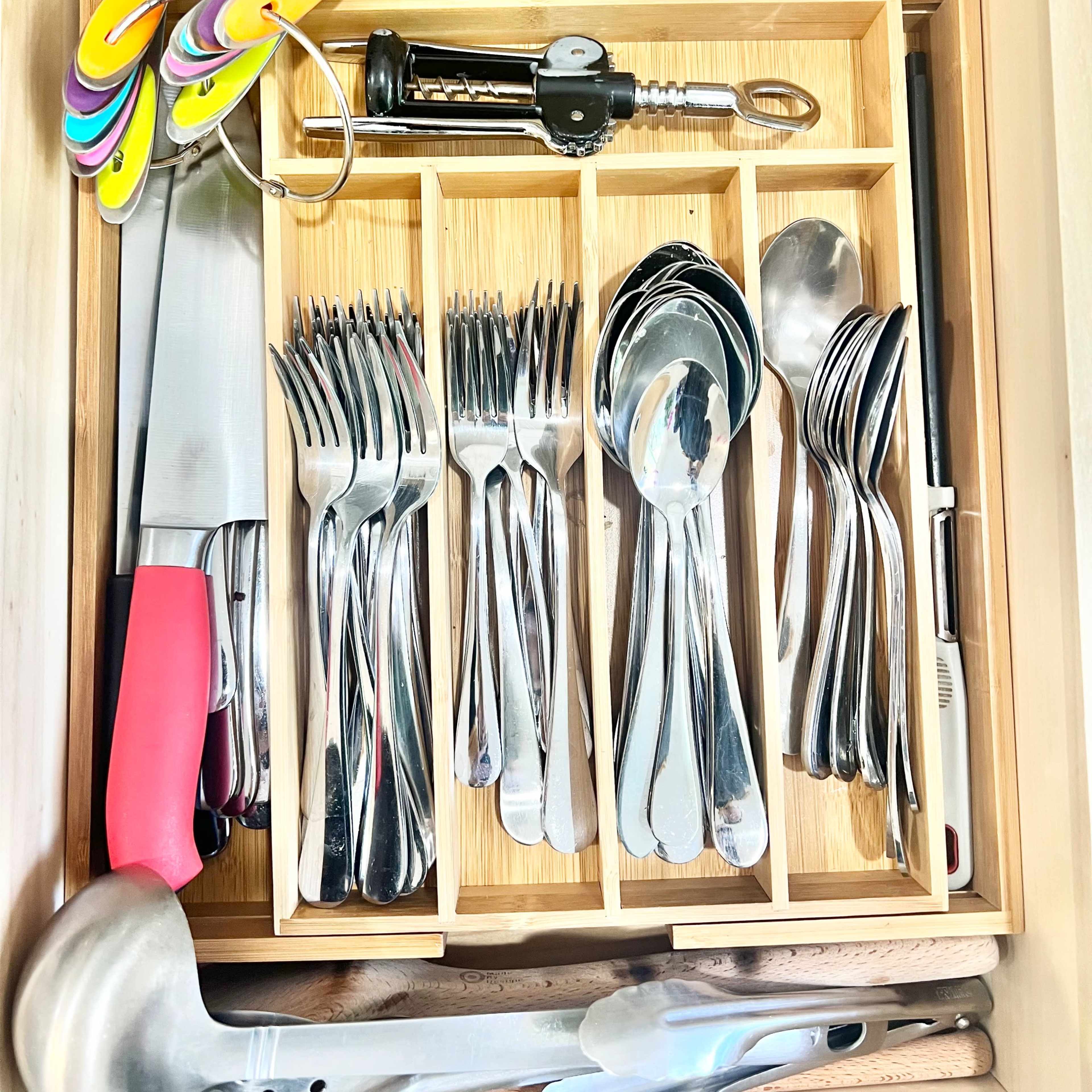 A wooden kitchen drawer contains an assortment of measuring cups, measuring spoons, knives, forks, spoons, and other utensils organized in compartments.