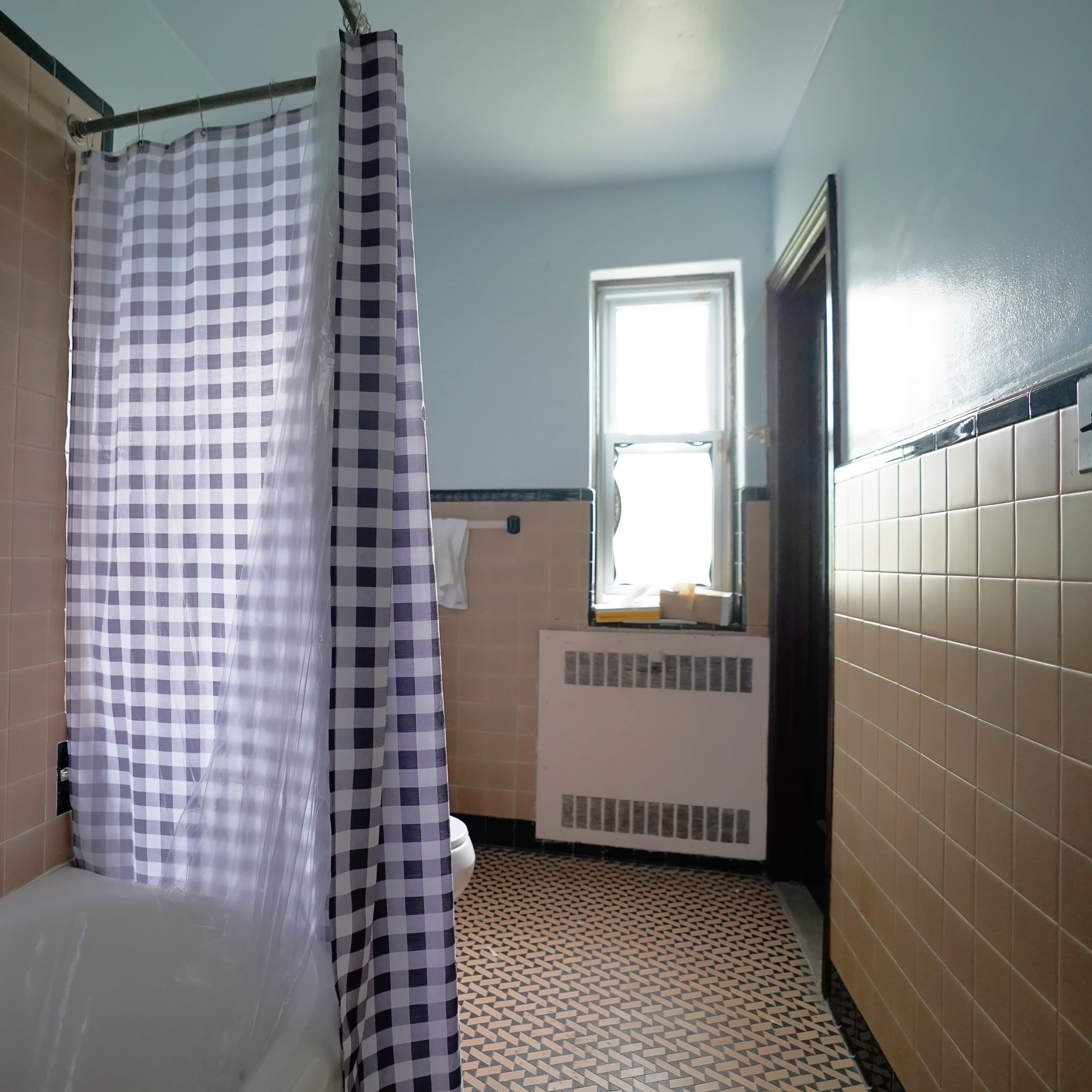 A bathroom featuring a bathtub with a checkered shower curtain, beige tiled walls, and a window allowing natural light.