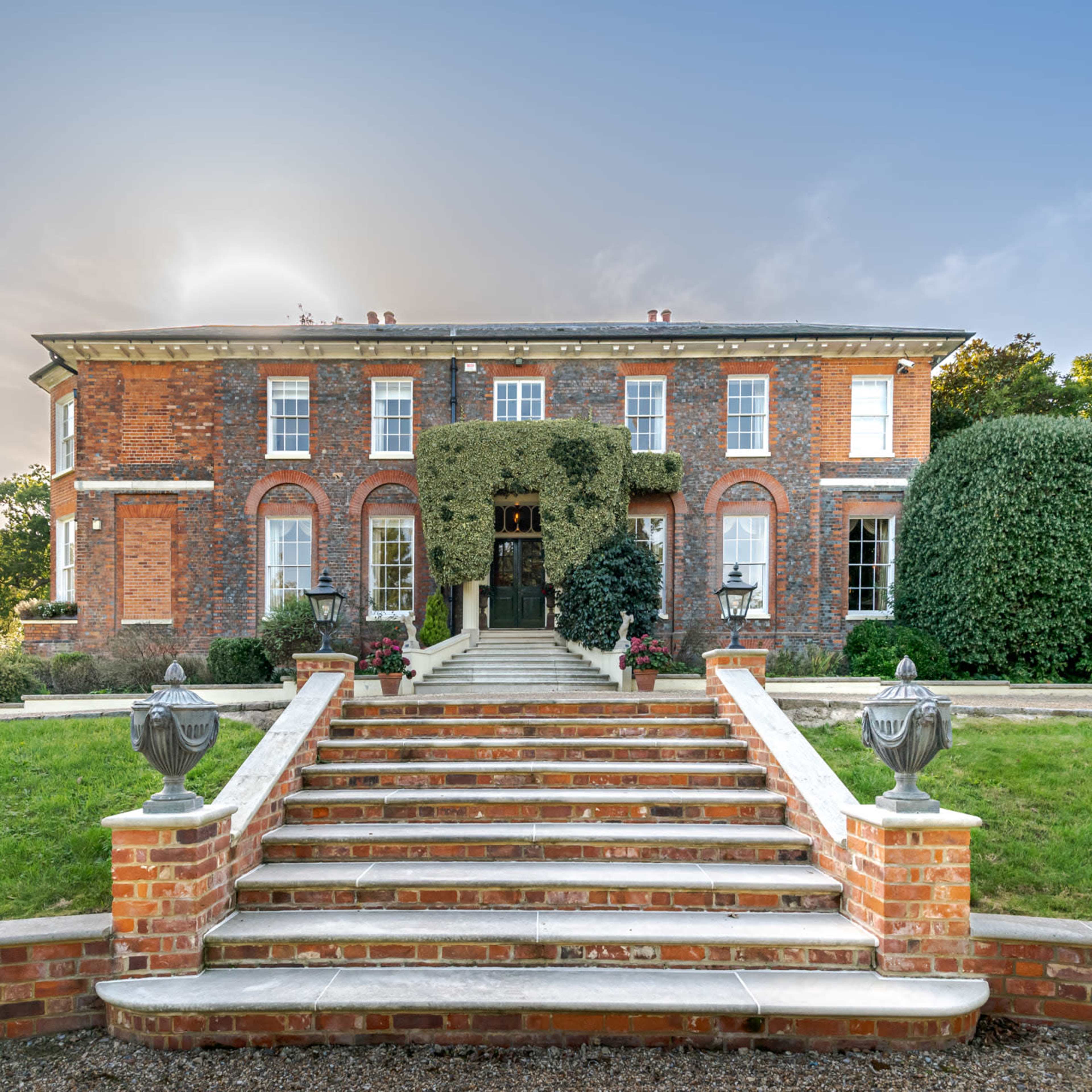 A grand brick house with a large staircase and symmetrical landscaping is set against a clear sky.