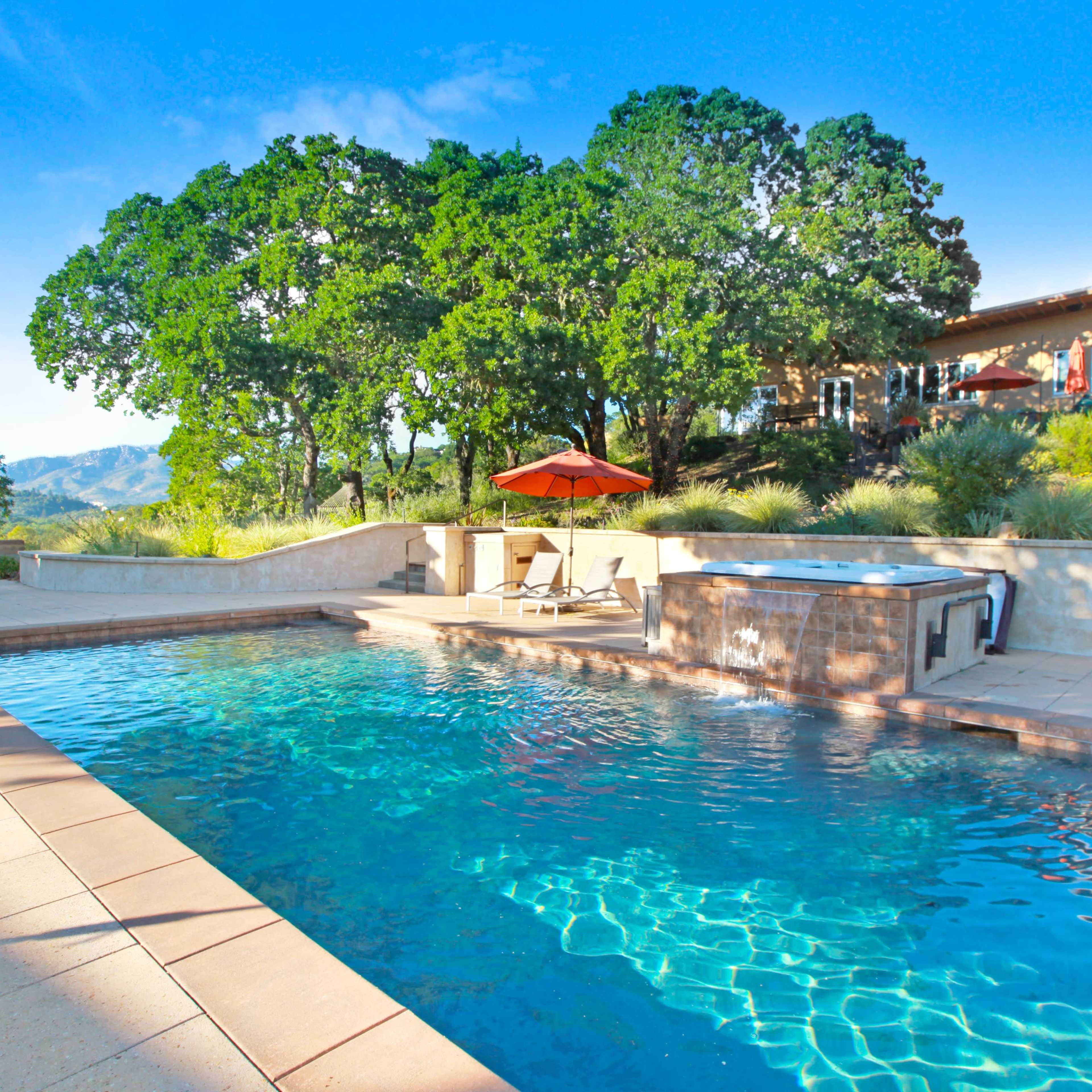 A swimming pool surrounded by a stone patio, with lounge chairs and an umbrella, set against a backdrop of trees and a hillside house.