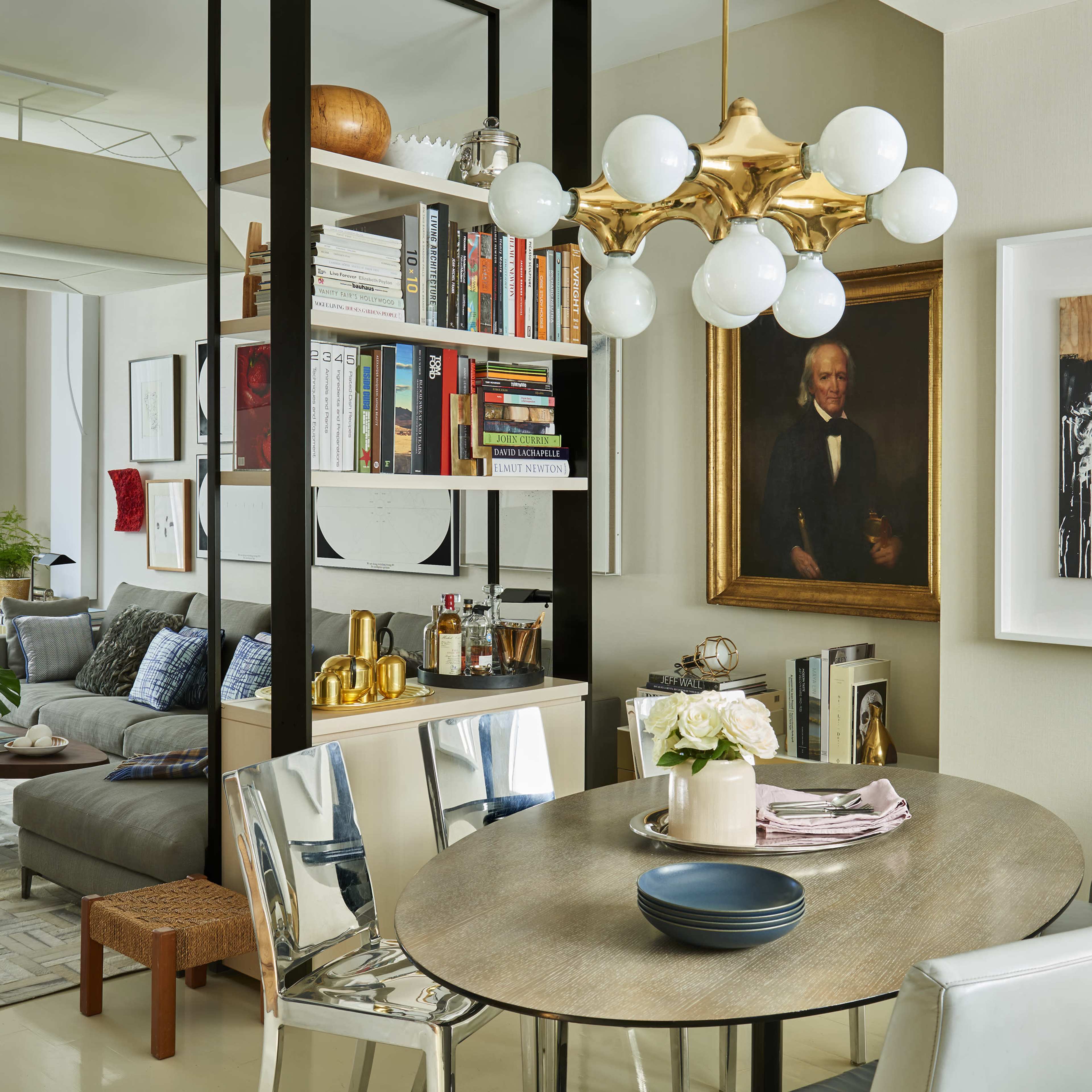 A modern living space with a dining area featuring a round table, a bookshelf, a framed portrait, and a comfortable seating area visible through an open layout.