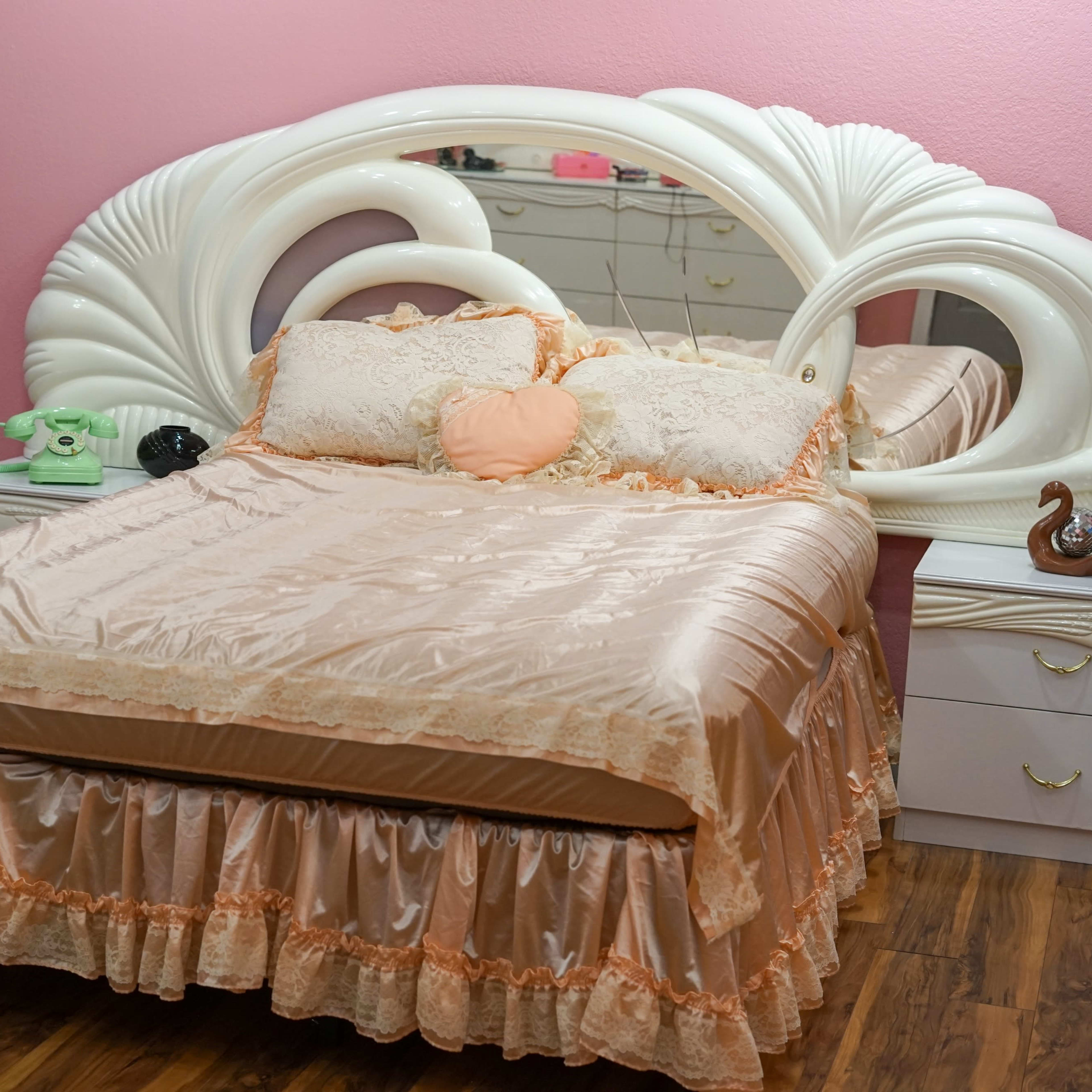 A styled bedroom with a bed featuring a decorative headboard, pastel bedding, and a vintage telephone on a bedside table against a pink wall.